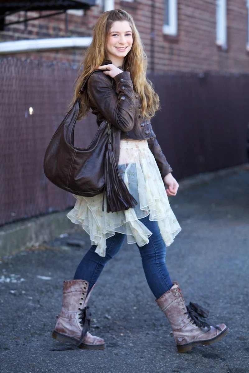 A young girl with long wavy hair, wearing a brown leather jacket, a layered cream-colored skirt, dark blue jeans, and lace-up combat boots, carrying a large dark brown handbag with tassels, standing outdoors on pavement near a brick building.