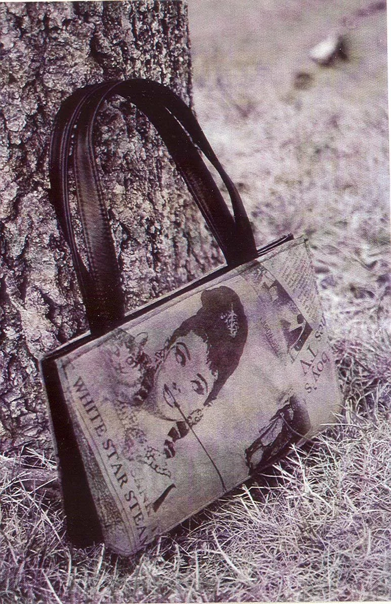 A vintage Glamour magazine tote bag leaning against a tree, with a portrait of a woman on the cover, placed on the ground with grass and a dirt path nearby.