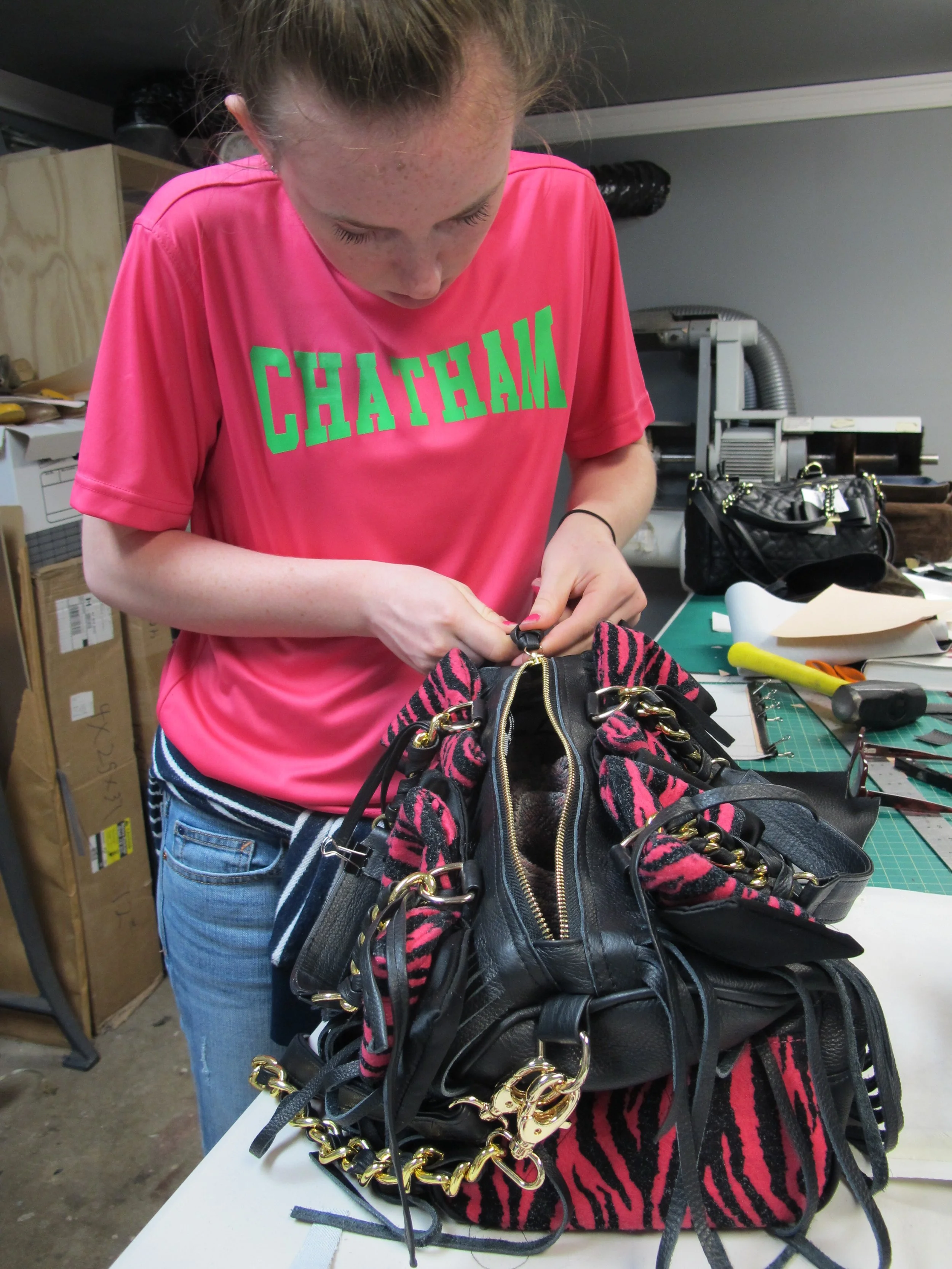 A woman working on a black handbag with pink and black striped fabric, gold hardware, and tools on a work table.