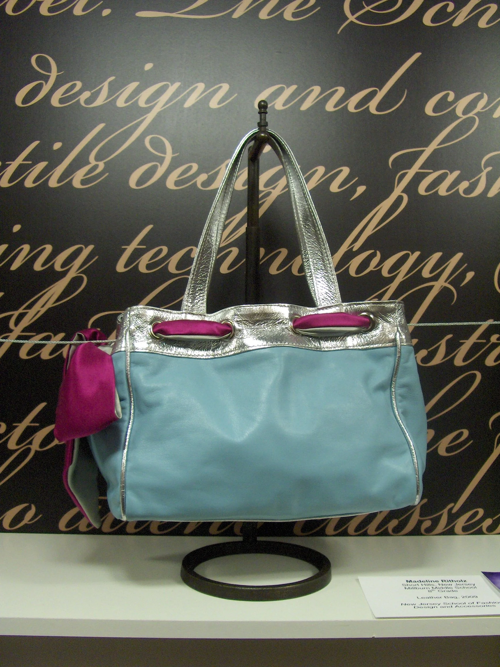 A light blue handbag with silver handles and trim, displayed on a stand with a purple accessory attached, against a background with cursive writing.