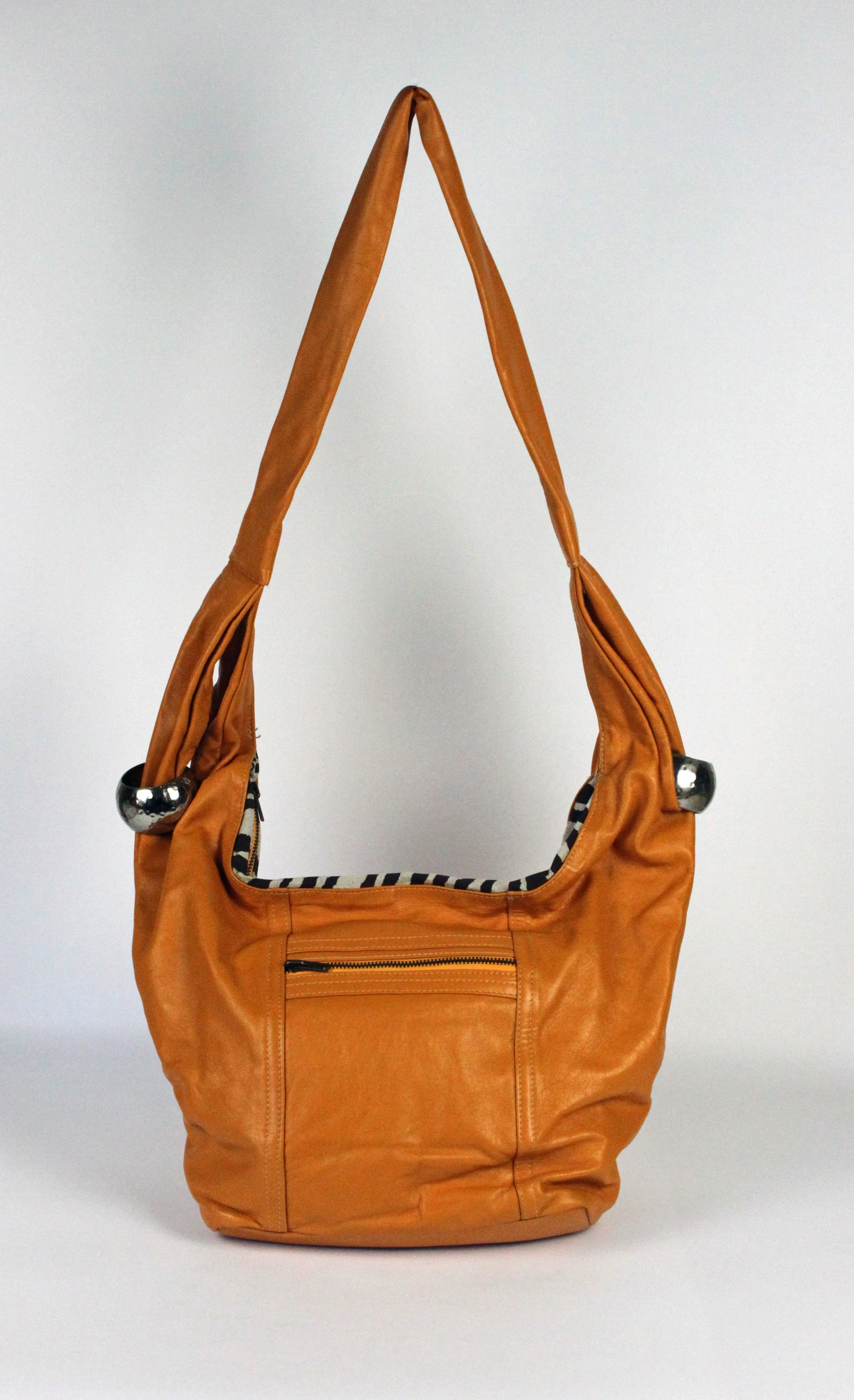 A brown leather shoulder bag with a zippered front pocket, silver hardware, and striped fabric lining, standing against a plain background.