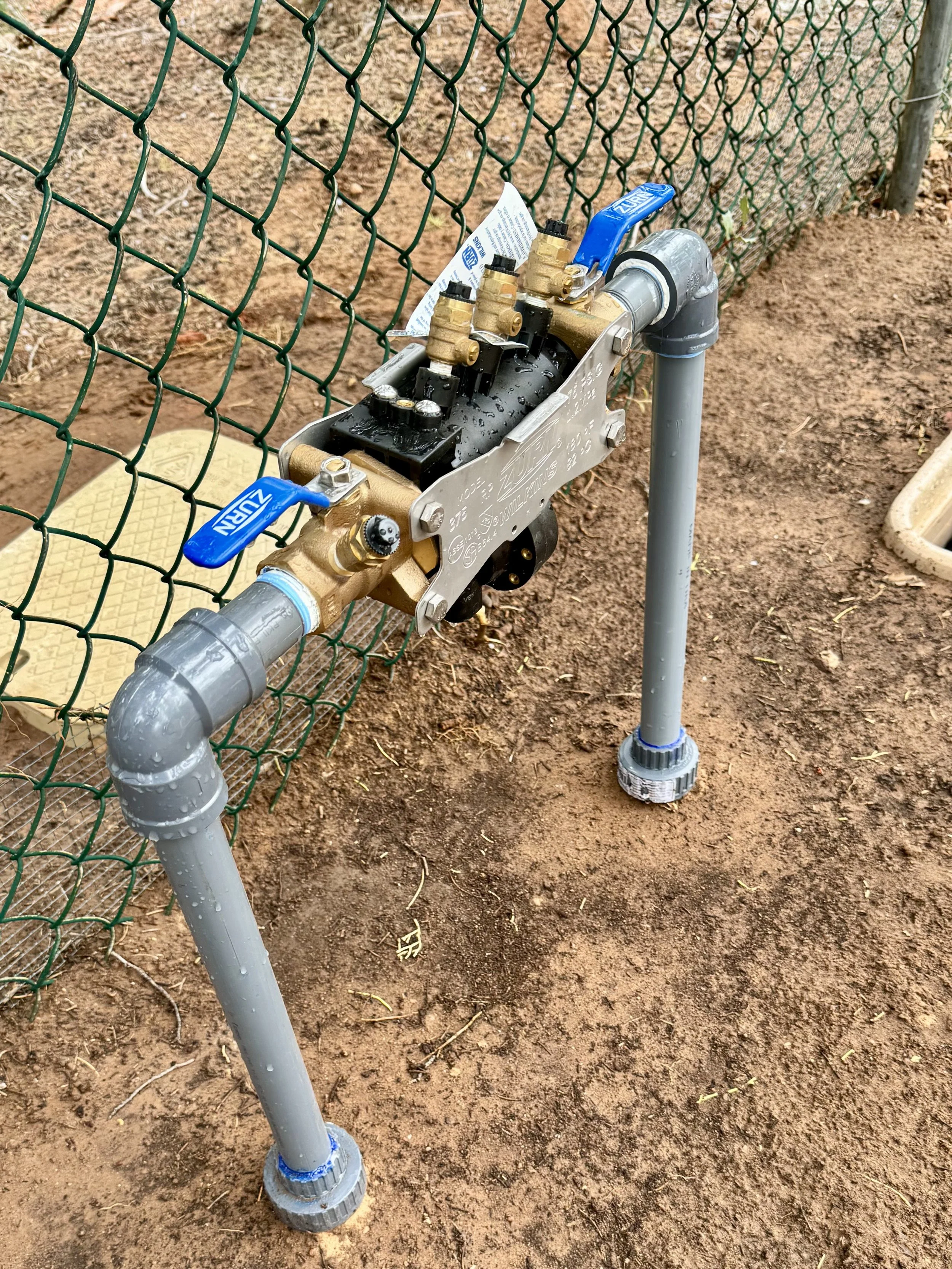 A backflow prevention  system behind a fence, with pipes and a valve handle, situated on dirt ground.