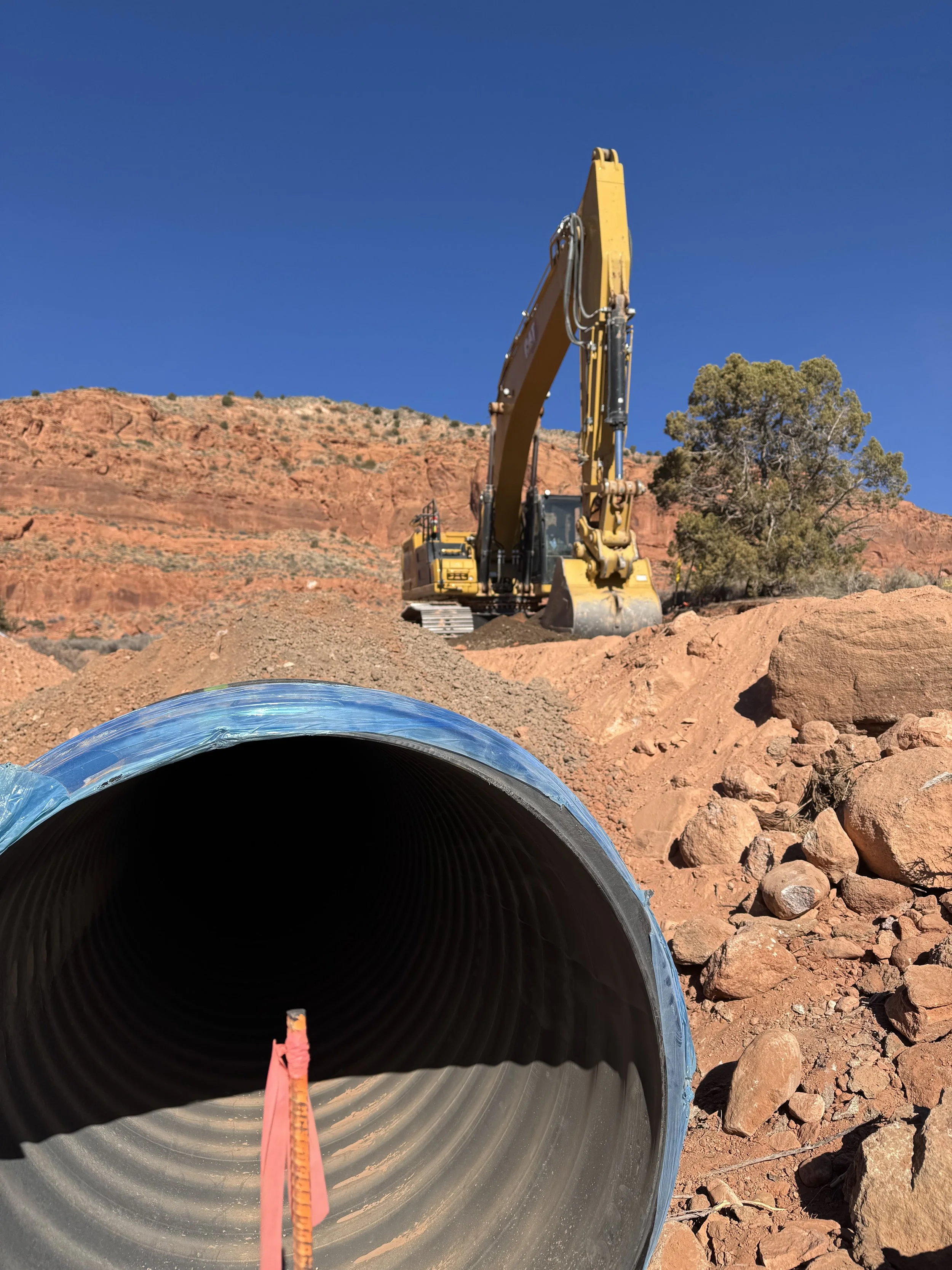 Excavator digging beside a large black pipeline in a red-rock desert landscape