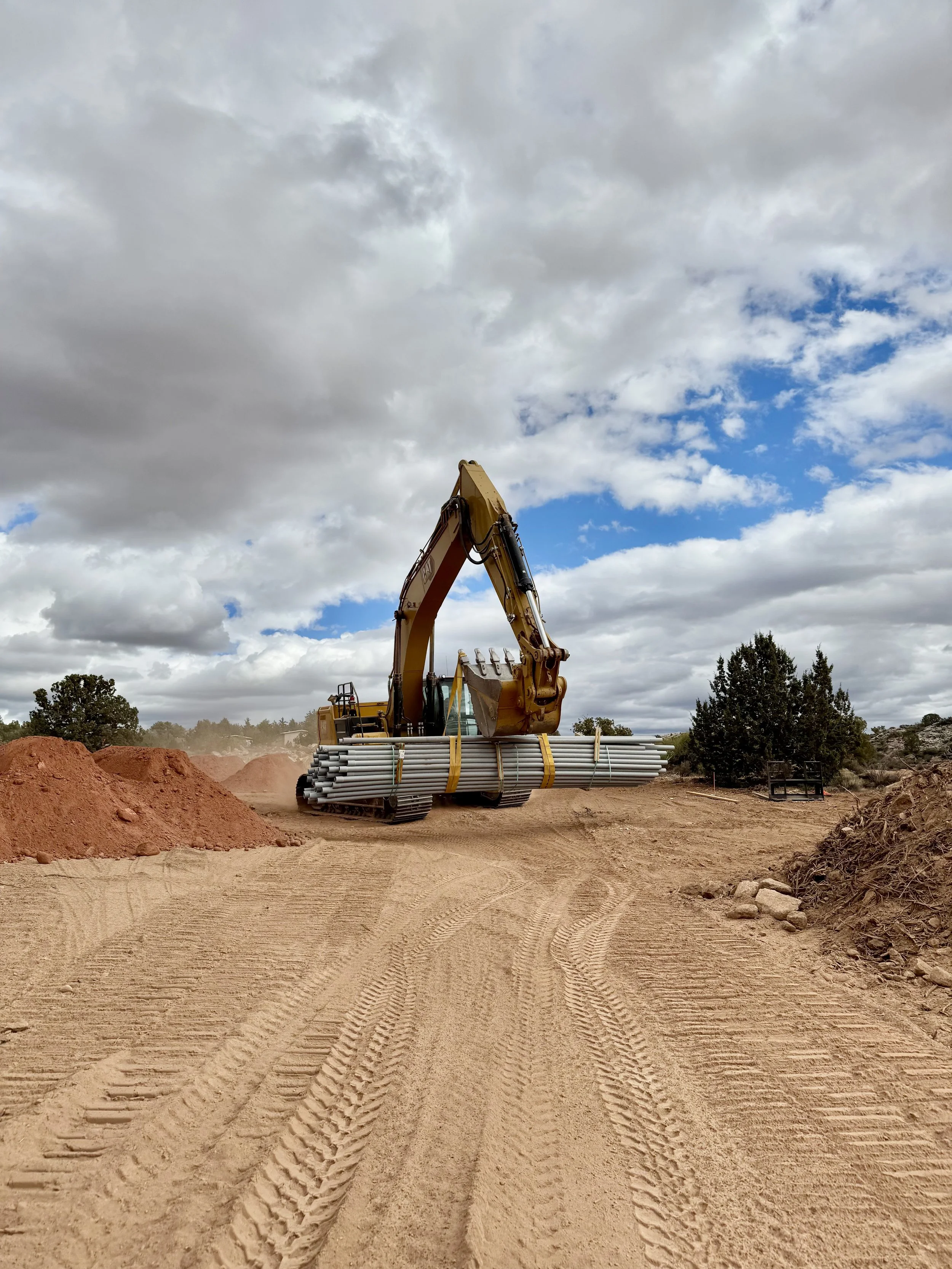 Yellow excavator carrying bundled pipes at a construction site beneath a cloudy blue sky