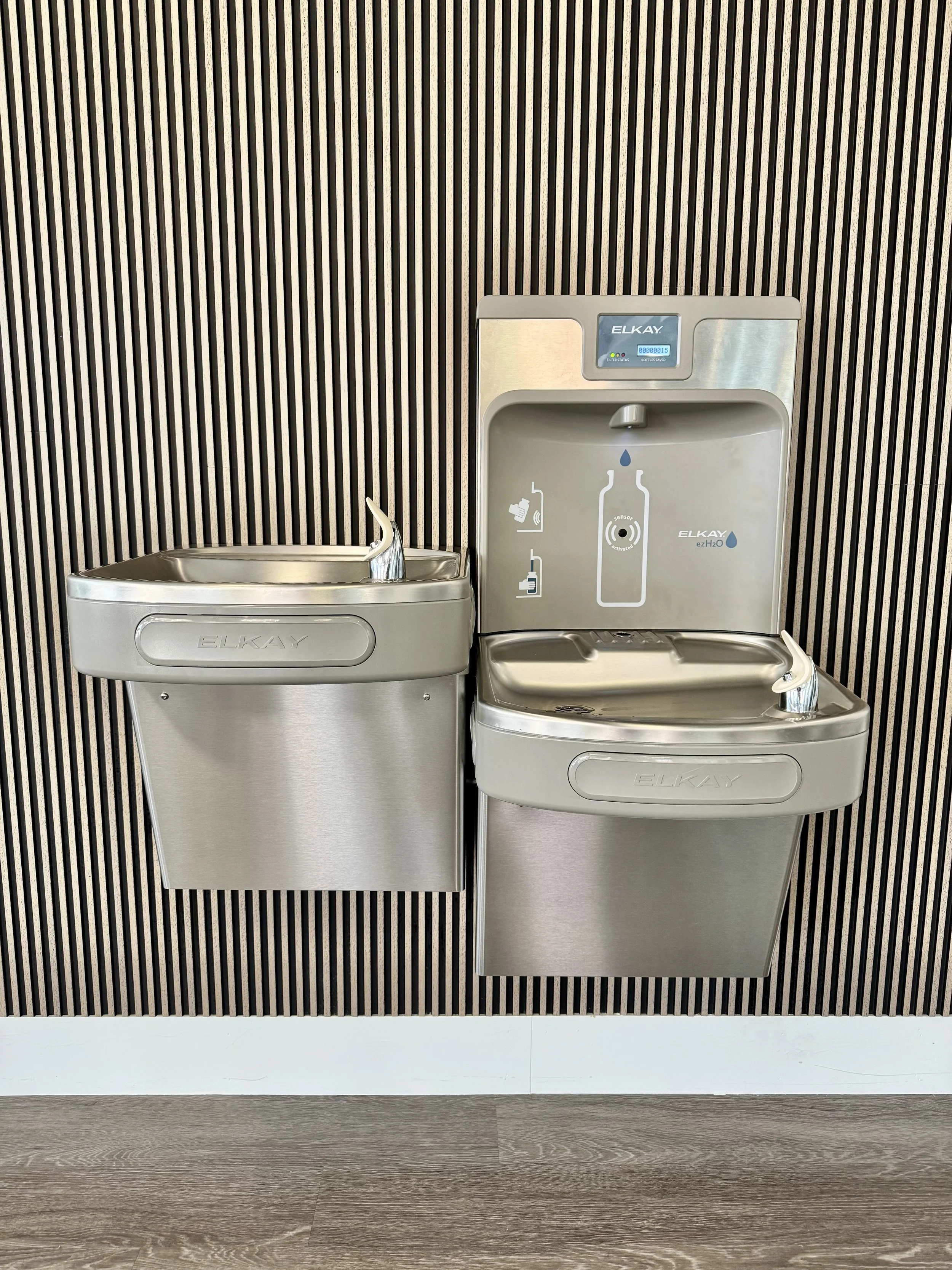 Two stainless steel water refill stations with black and cream striped background and wood-look flooring.