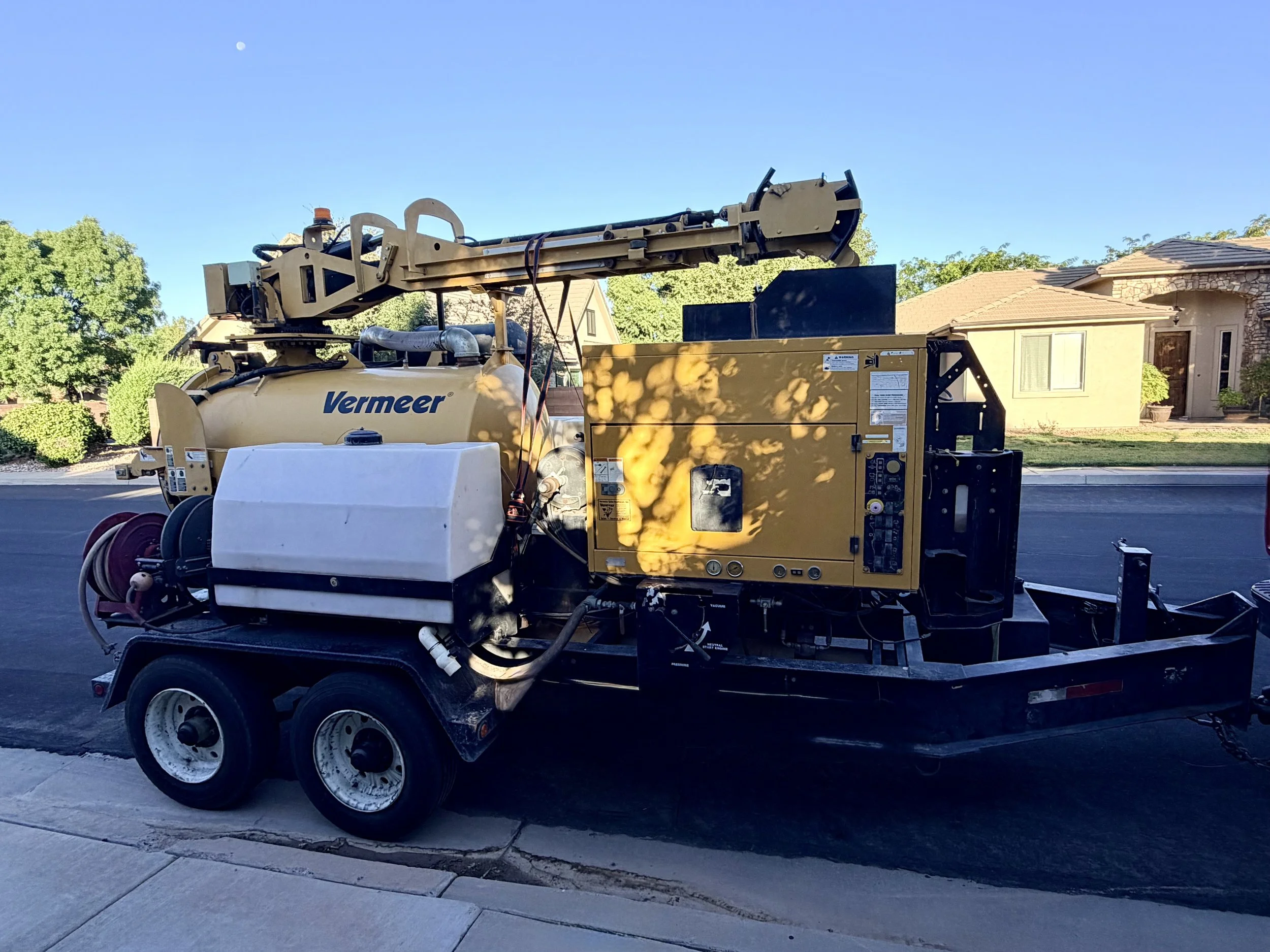 A beige Vermeer wood chipper machine on a trailer parked on a residential street with houses and trees in the background, and a clear blue sky overhead.