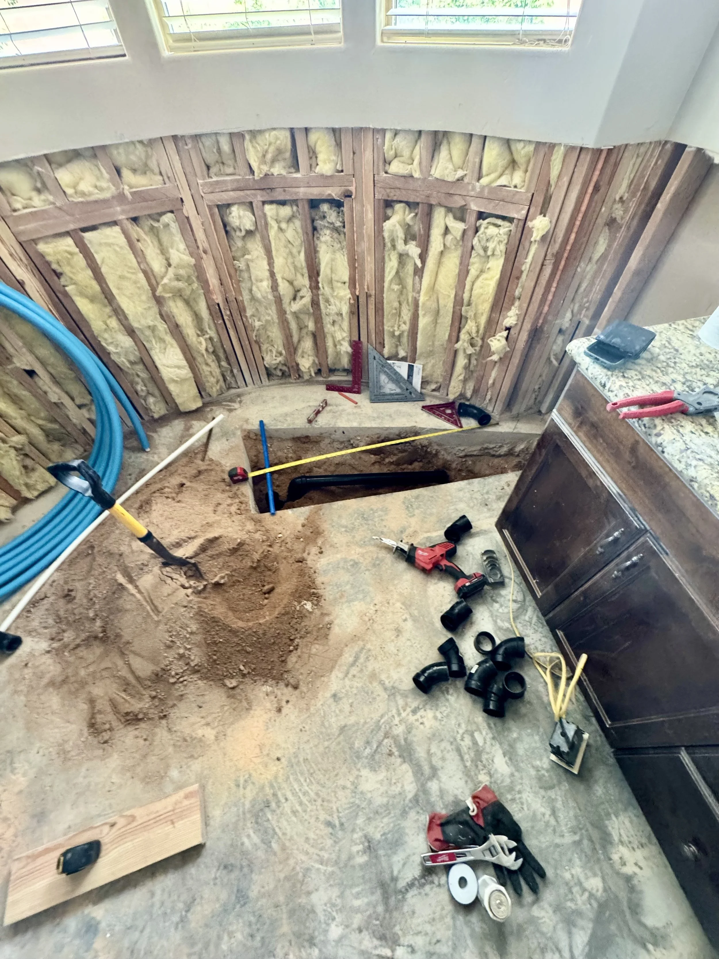 Room undergoing renovation, with exposed wall studs and insulation. Tools and materials are scattered on the floor and surface, including a tape measure, saw, drill, and various pipes and fittings. A countertop with a phone and pliers is near the wall. Two windows are at the top of the image.