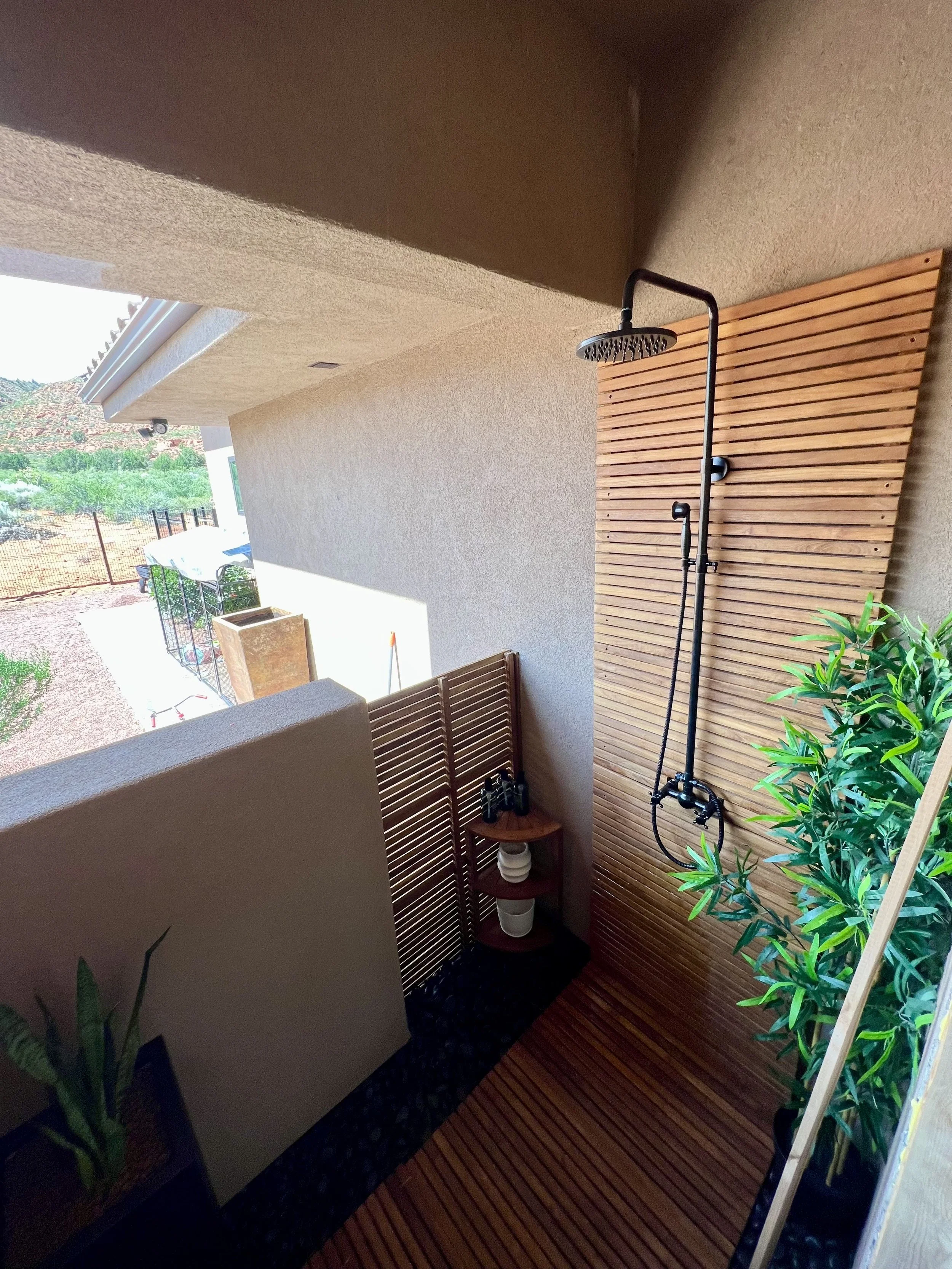 Outdoor patio area with a shower featuring a rainfall showerhead mounted on a wooden slat wall, potted plants, and lounge chairs.