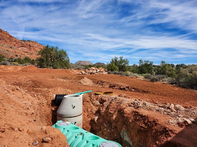 A construction site in a desert landscape with red soil, a pipe extending from a drainage pipe, and a partly cloudy sky.
