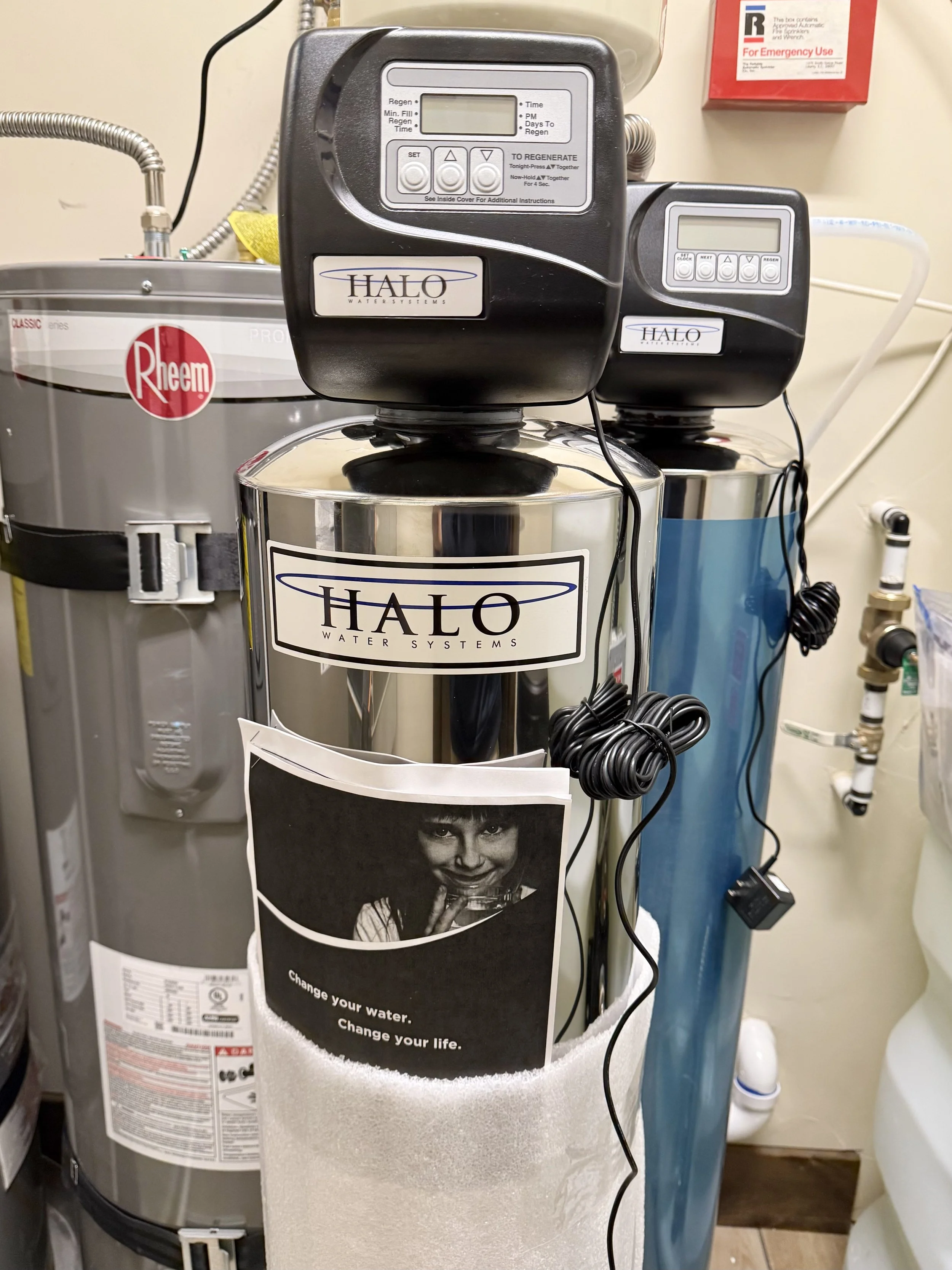 Two HALO Water Systems water filters connected to a water line, with control panels on top, in a utility room. A Rheem water heater is visible on the left side, and various pipes and a red fire safety box are seen in the background.