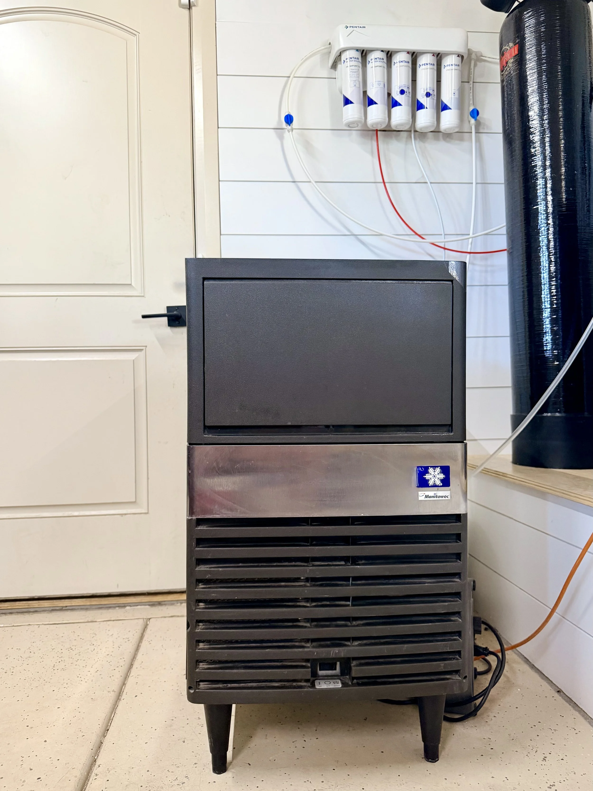 A water filtration system with five filter cartridges mounted on a wall, a black water tank, a black metal appliance with vent slats, and an orange tube on a wooden platform against a white paneled wall.
