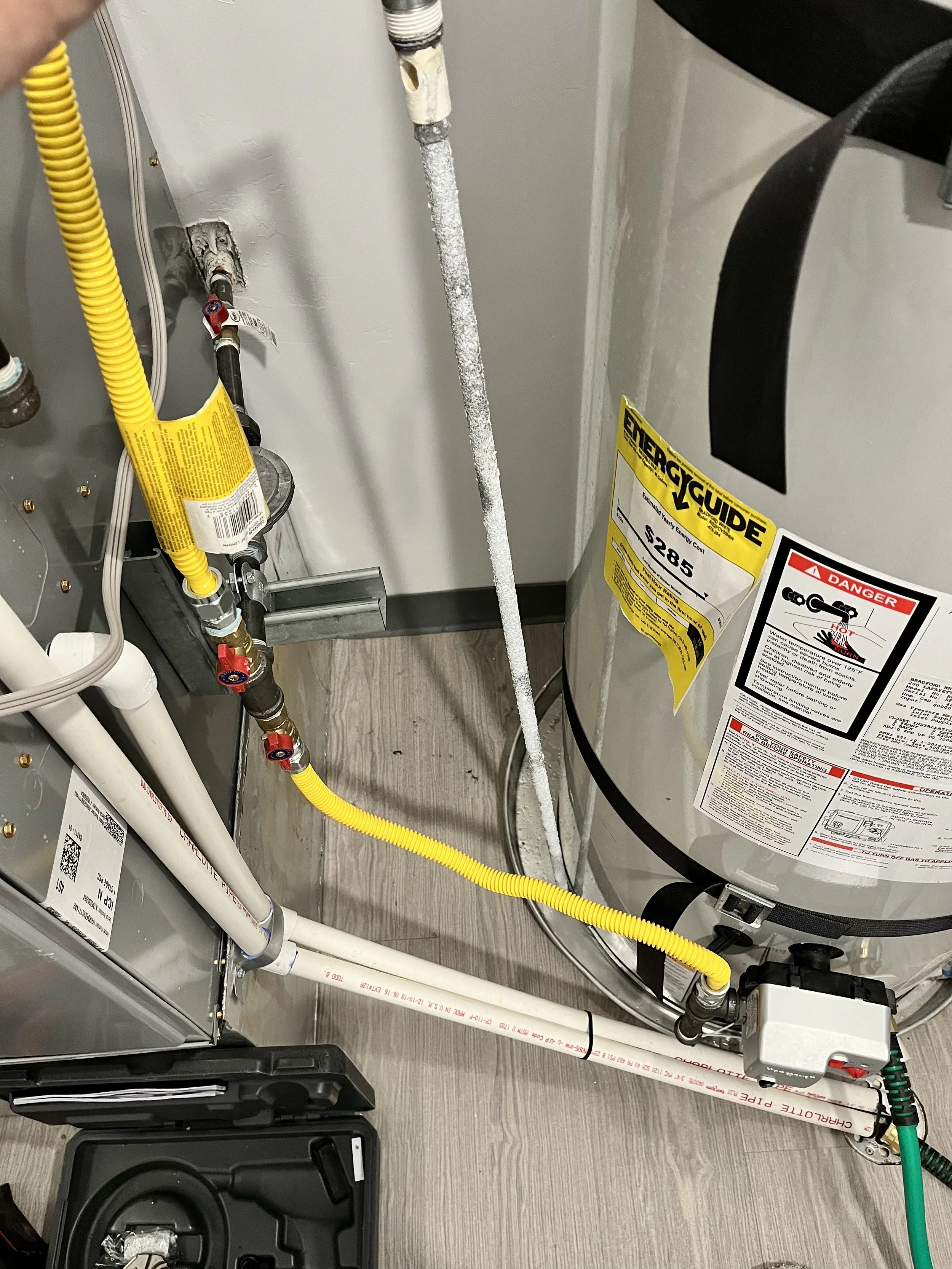 Interior of a utility or water heater room showing pipes, valves, and a water heater unit with labels and warning stickers. There are various colored pipes, including yellow, white, and black, connected to the equipment.