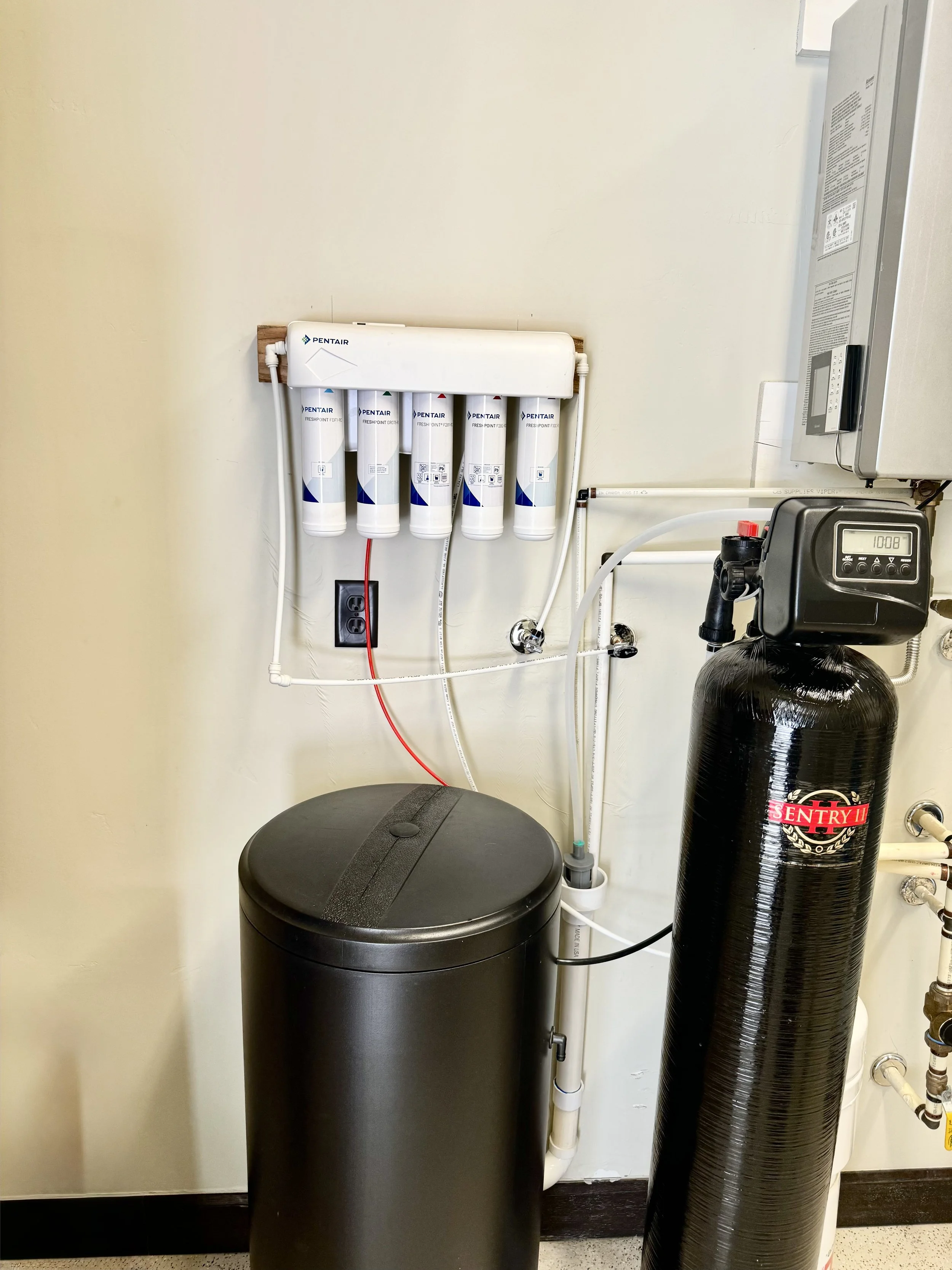 A water filtration system with five filter cartridges mounted on a wall above a large black water softener tank in a utility room.