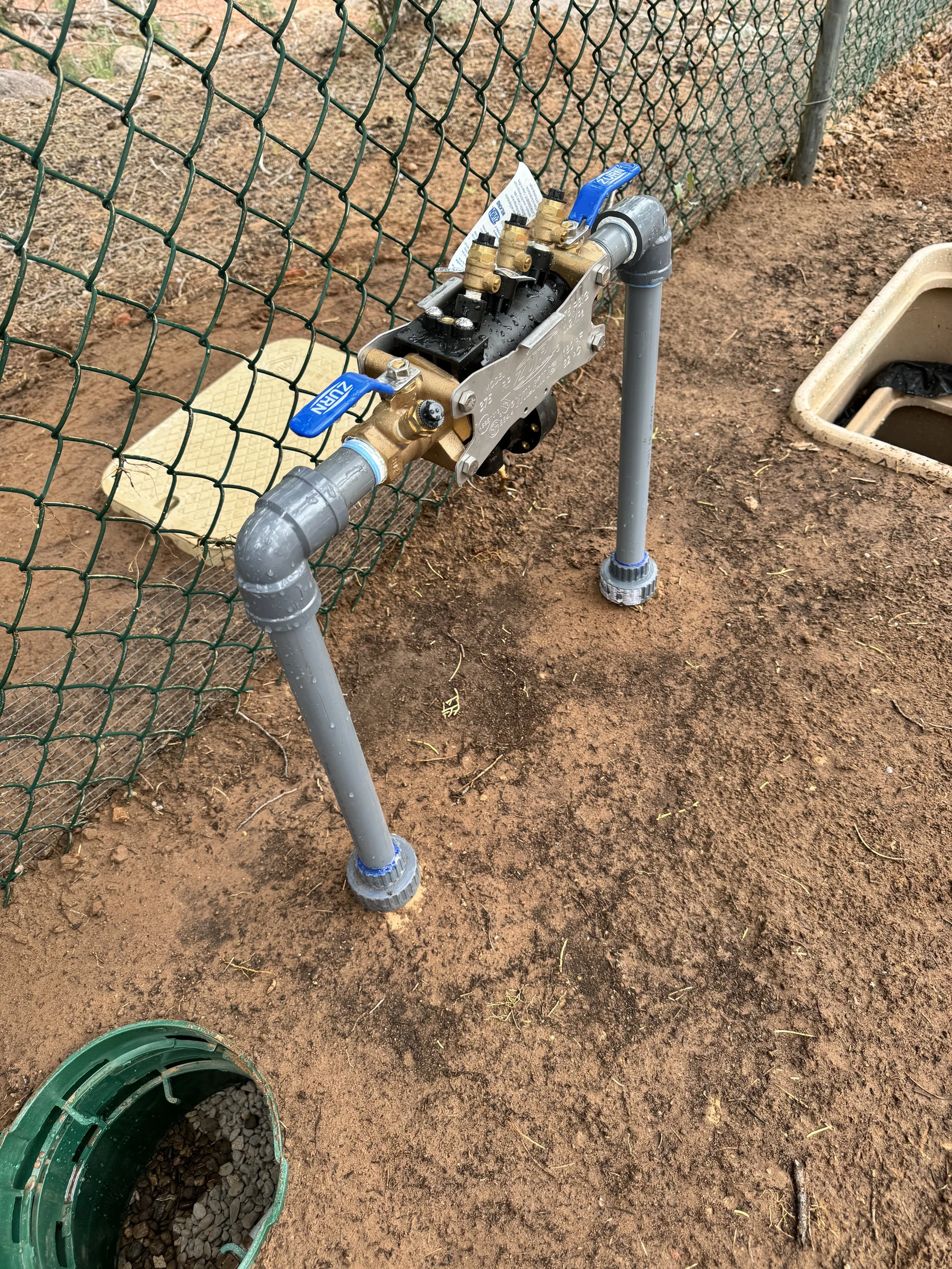 An irrigation system with a water valve attached to a pressure regulator and a pipe, installed outdoors on the ground near a chain-link fence. There is a green container filled with gravel in the bottom left corner. The ground appears to be dirt.