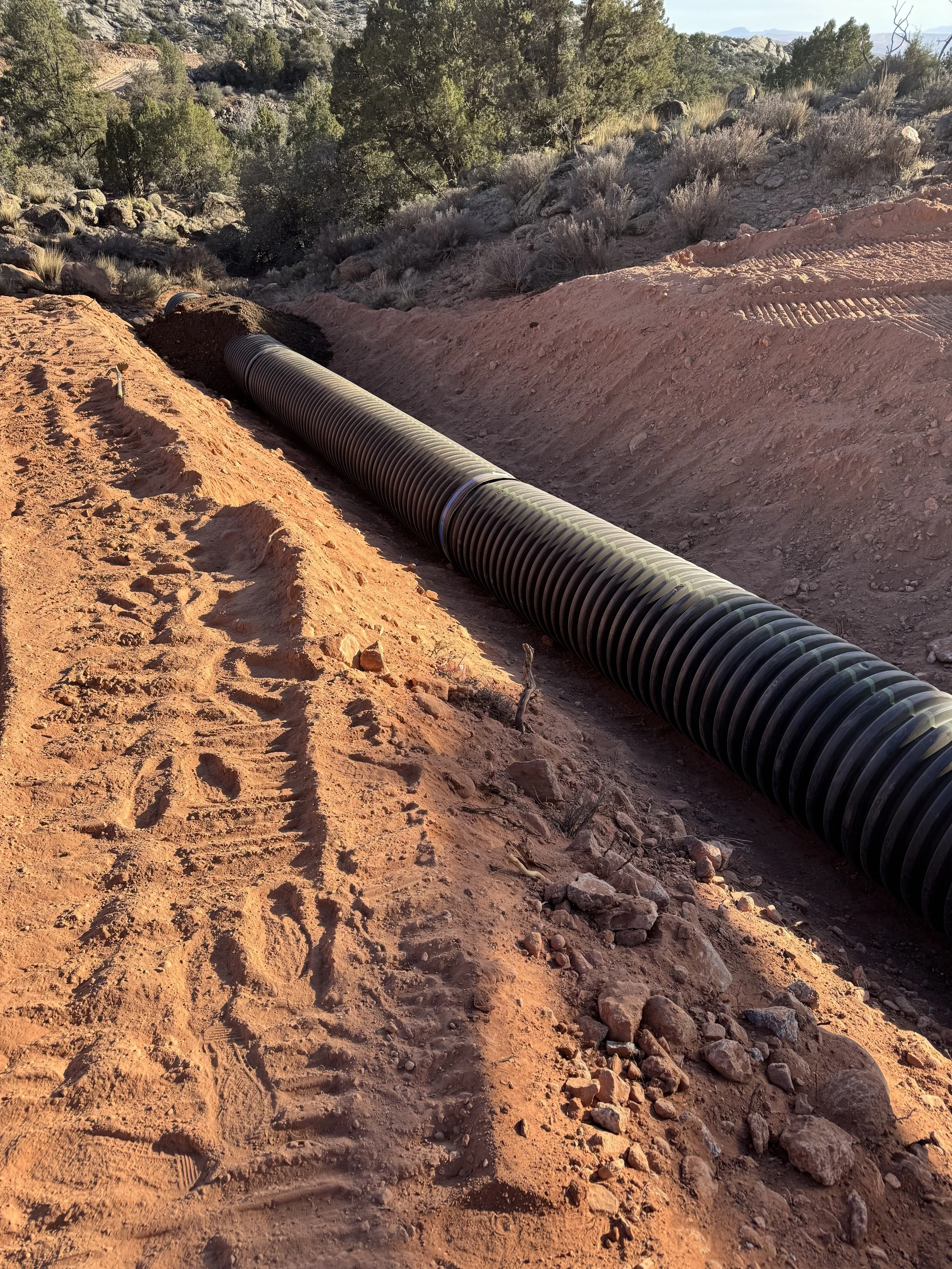 Large corrugated drainage pipe installed in a red dirt trench amid rocky desert vegetation and distant mountains