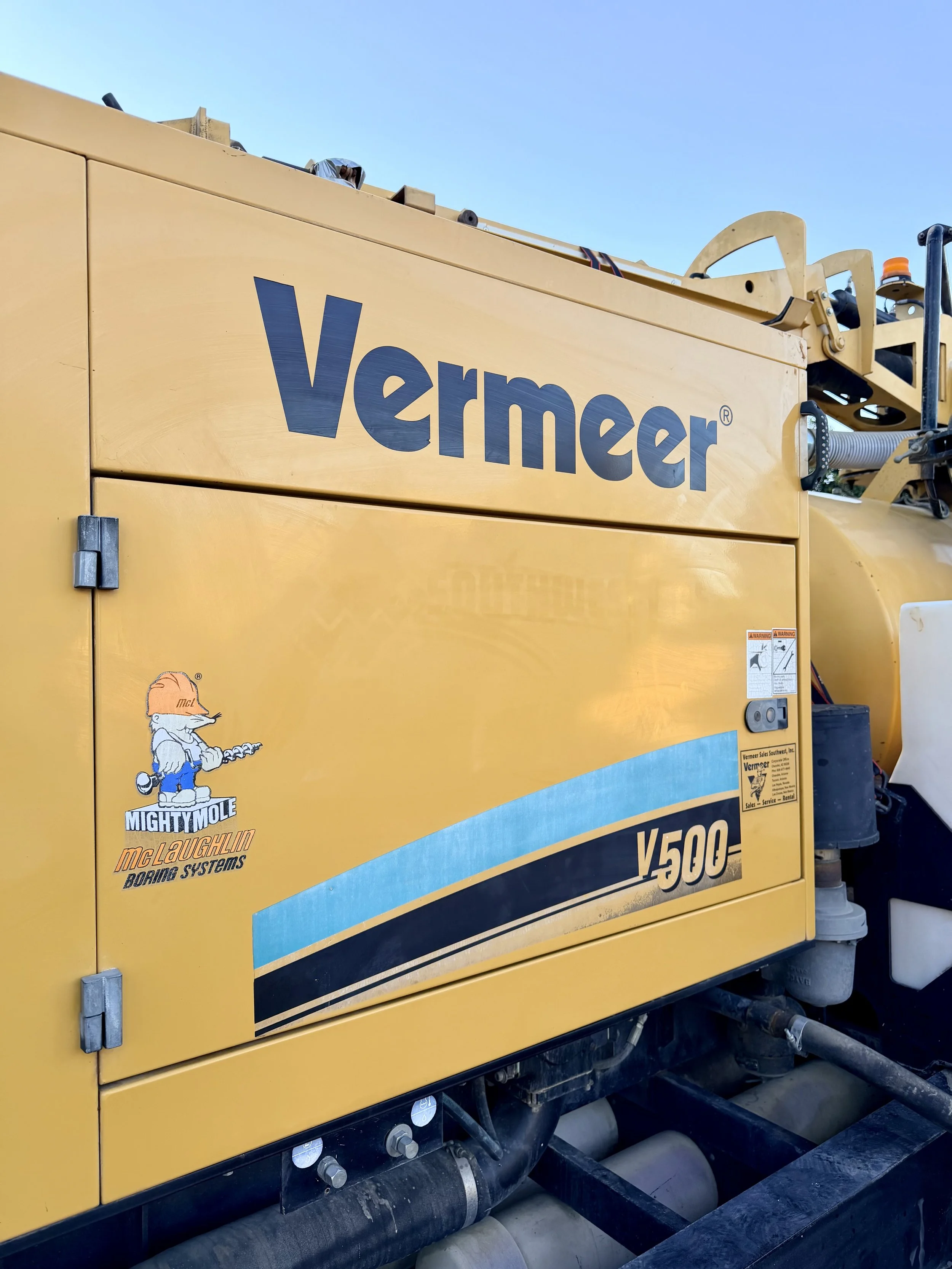 Close-up of a yellow Vermeer V500 hydrocac trailer mounted hydro vaccuum industrial machine with logos and stickers, including a cartoon mole mascot. The machine is on a construction site with visible pipes and a blue sky in the background.