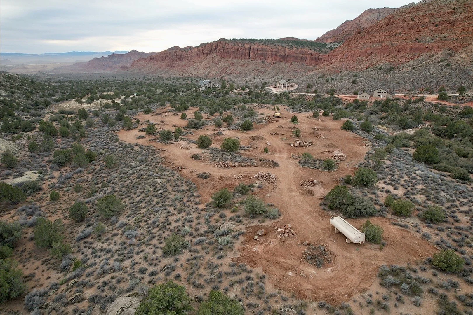 Aerial view of a red-rock desert landscape with homes, dirt roads, shrubs, and an empty trailer beneath rugged sandstone cliffs