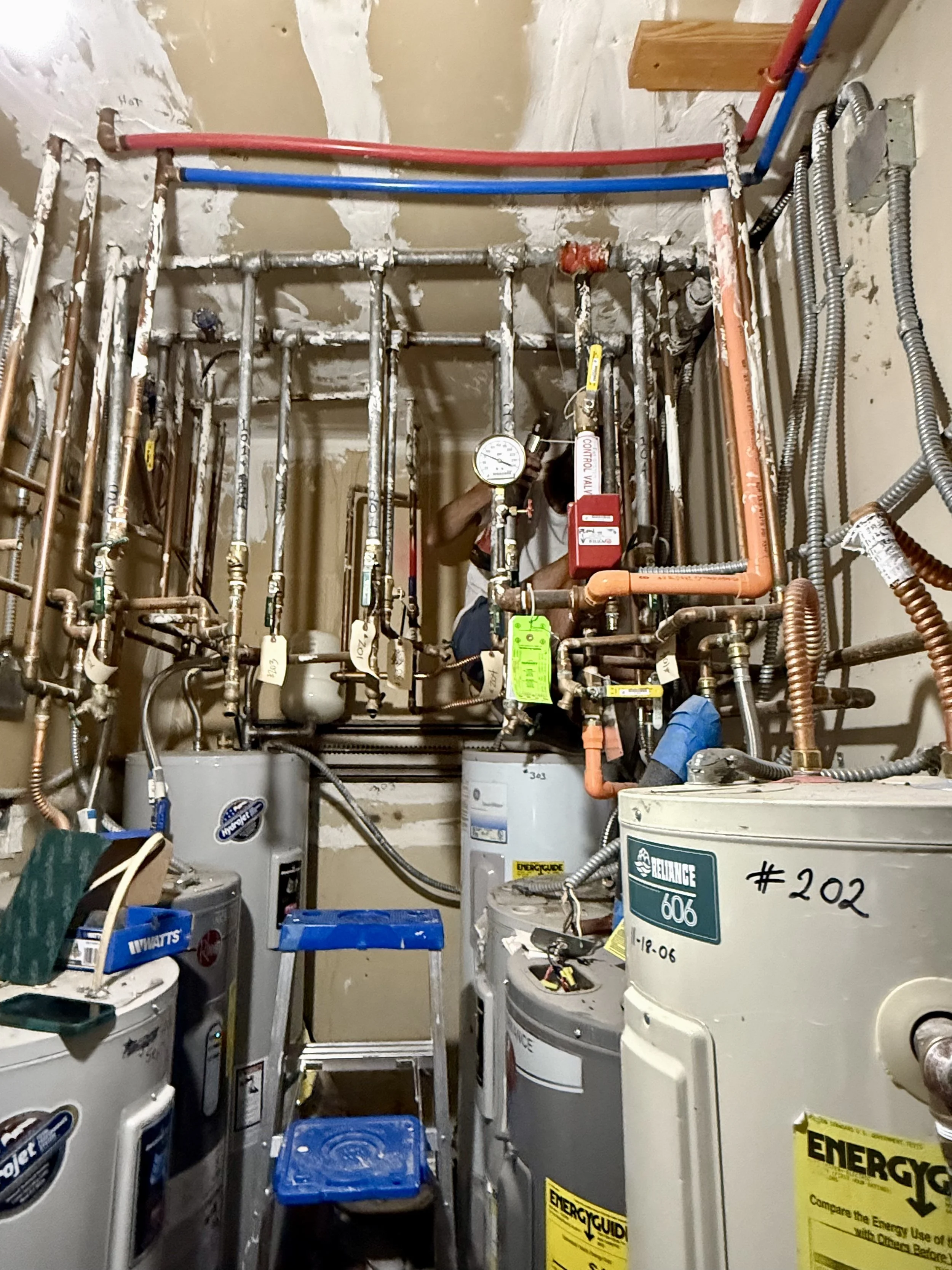 A utility room with exposed copper and plastic water pipes, a water heater, and a pressure gauge, with a lintel and tools in the background.