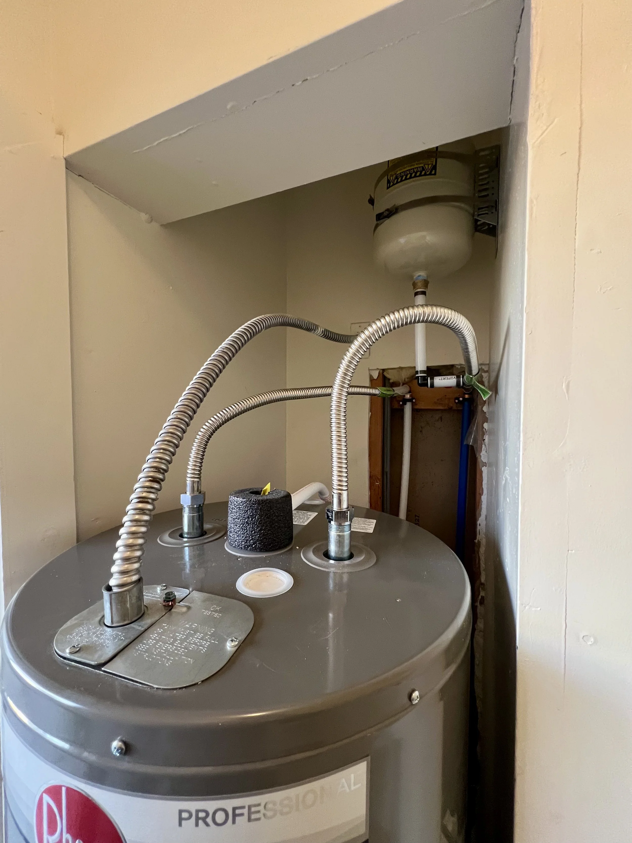 A water heater with electrical wires and plumbing pipes attached, installed in a wall cavity.
