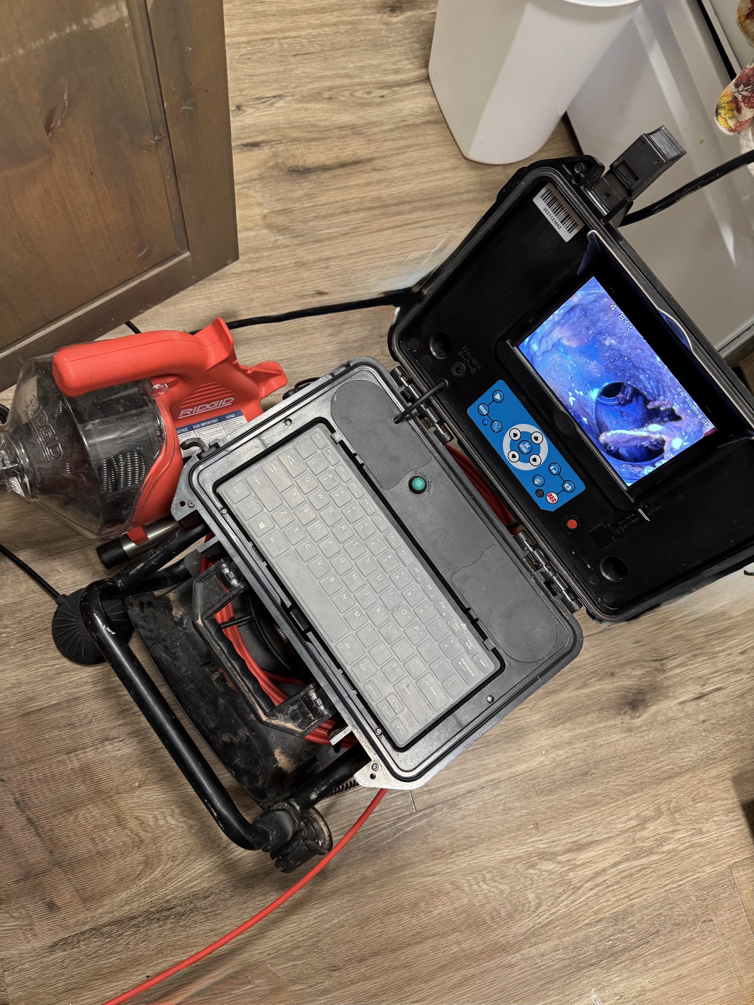 A sewer camera and leak locate device with a screen inside of the pipes, attached to a black metal stand with a red and black cable connected. Part of a wooden floor and a shoe cabinet are visible.
