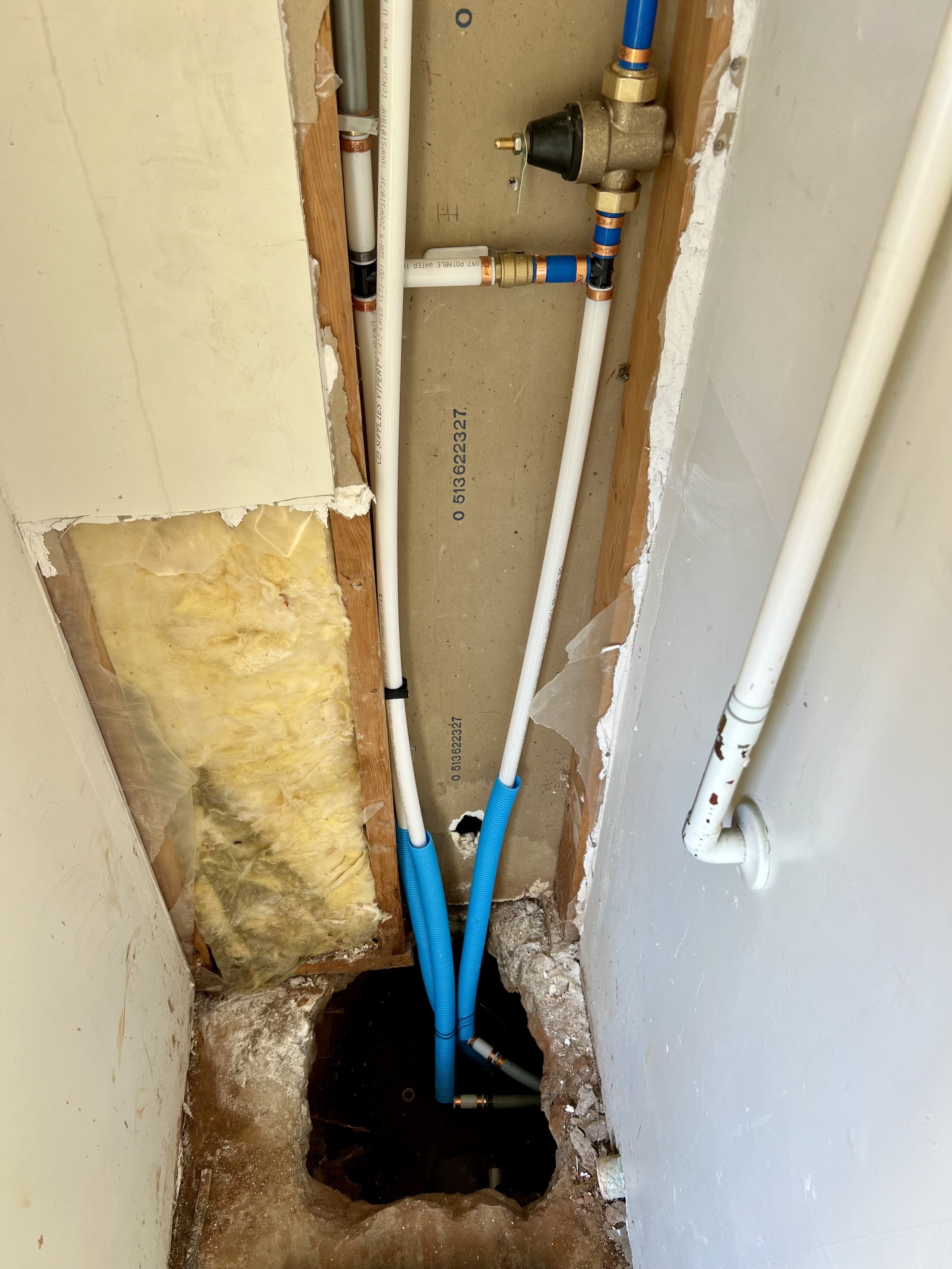 Plumbing pipes and a water pressure regulator in an unfinished wall opening, with insulation and drywall edges visible.