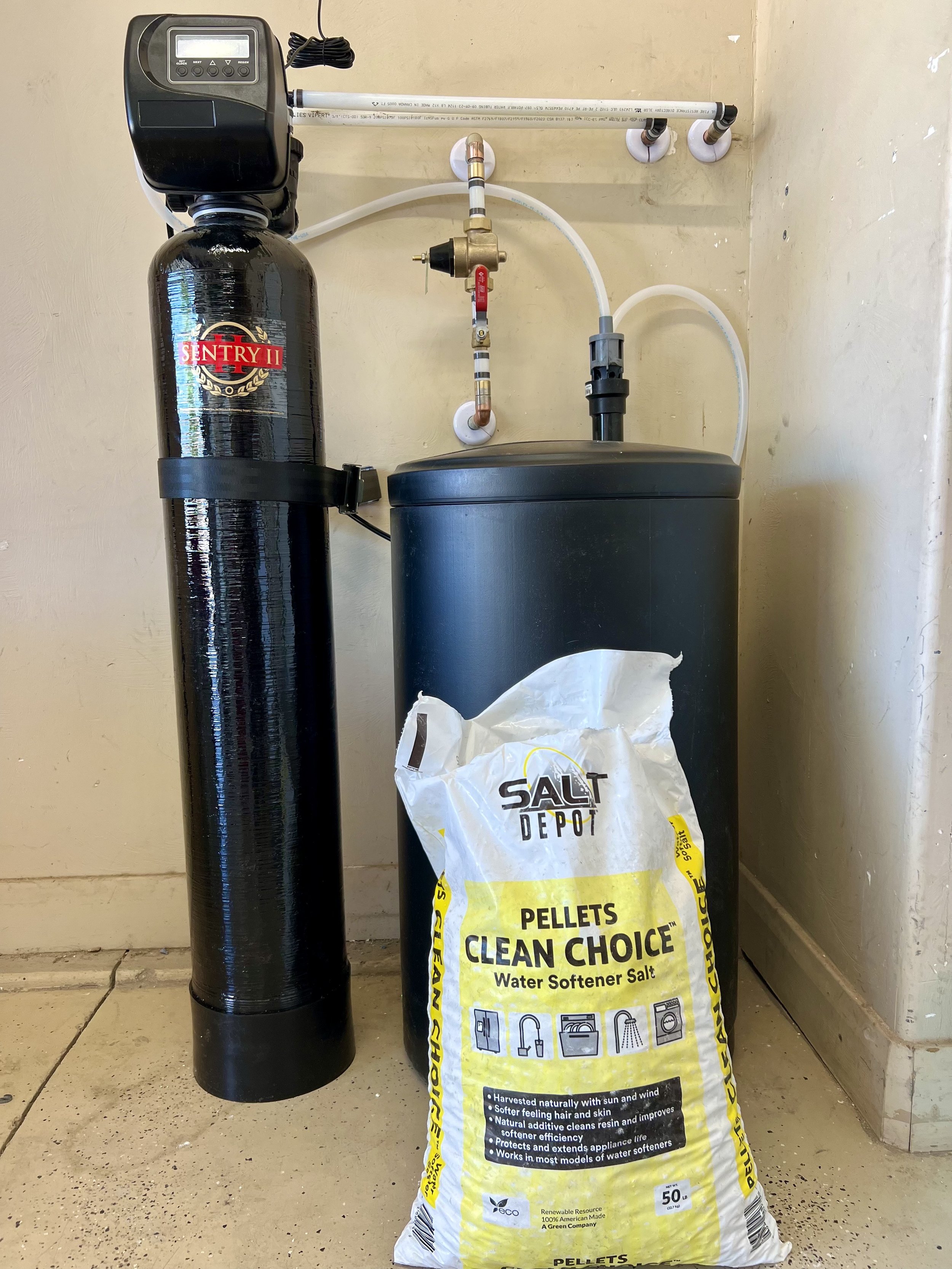 A water softener salt pellet bag in front of a water softener system with a black control valve and a black tank on a beige tiled floor against a beige wall.