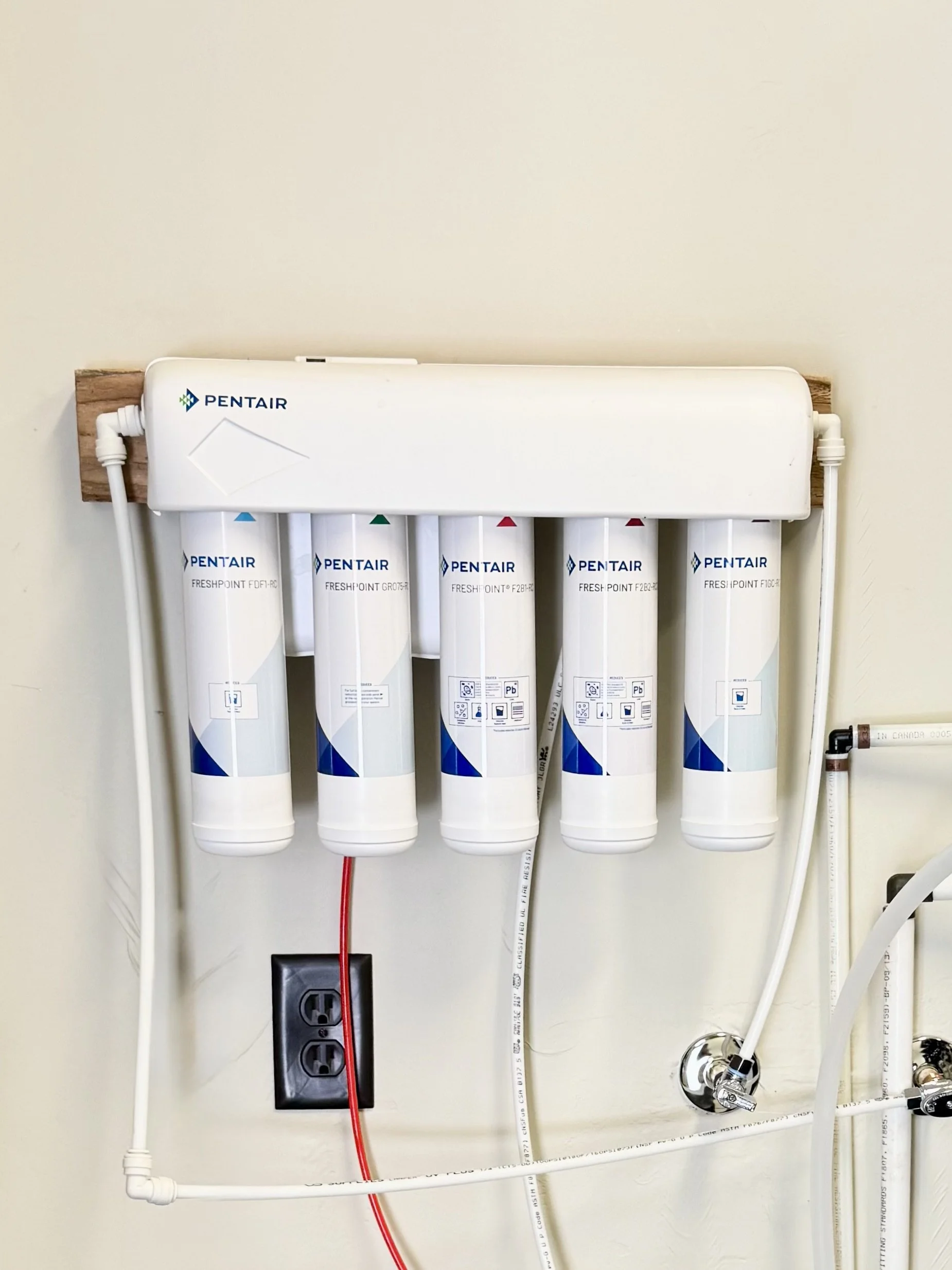 Water filtration system with five filters mounted on a wall, connected to white pipes and an electrical outlet below.