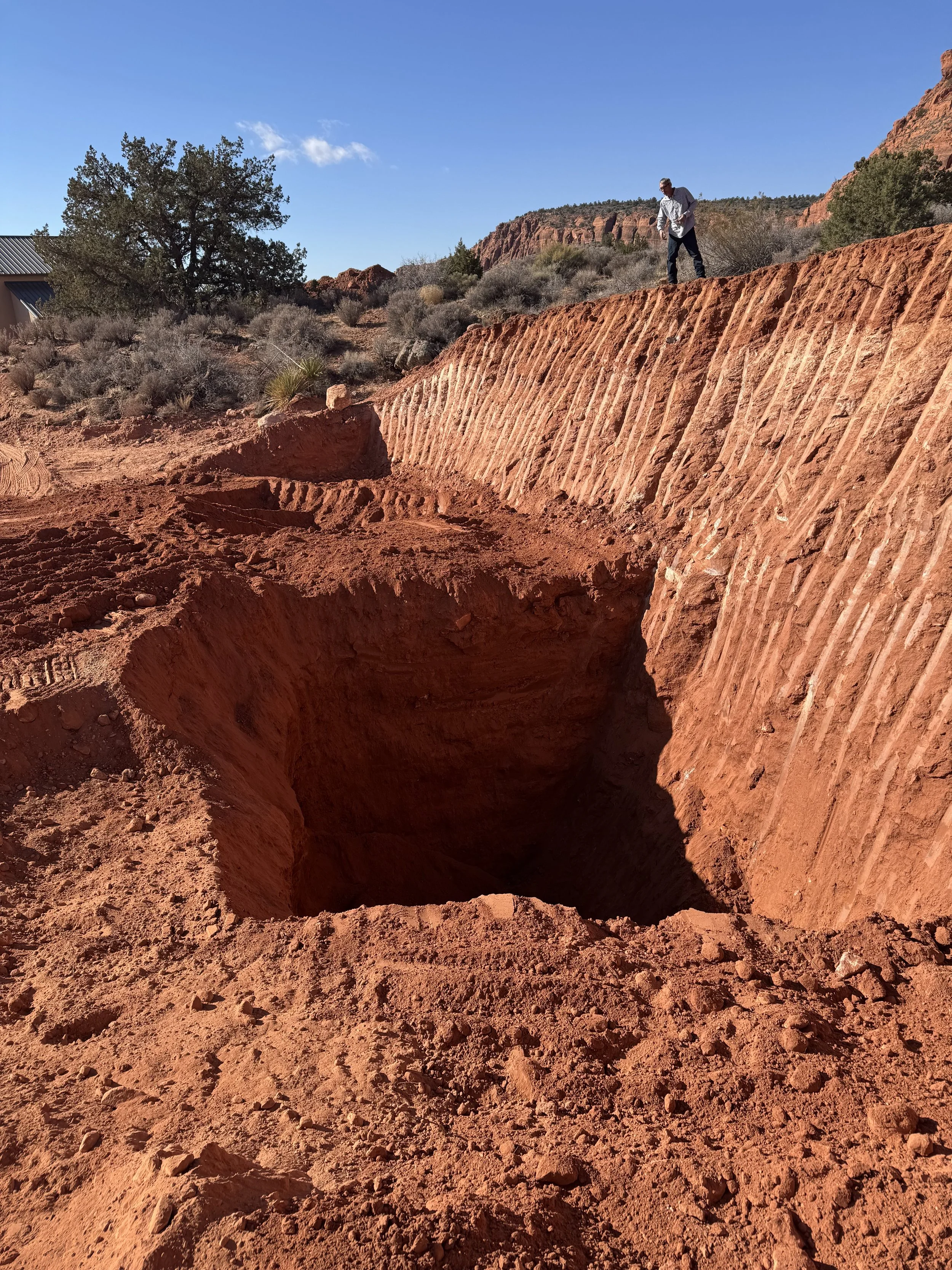 Person standing beside a deep trench in red desert soil, with red rock cliffs, shrubs, and blue sky