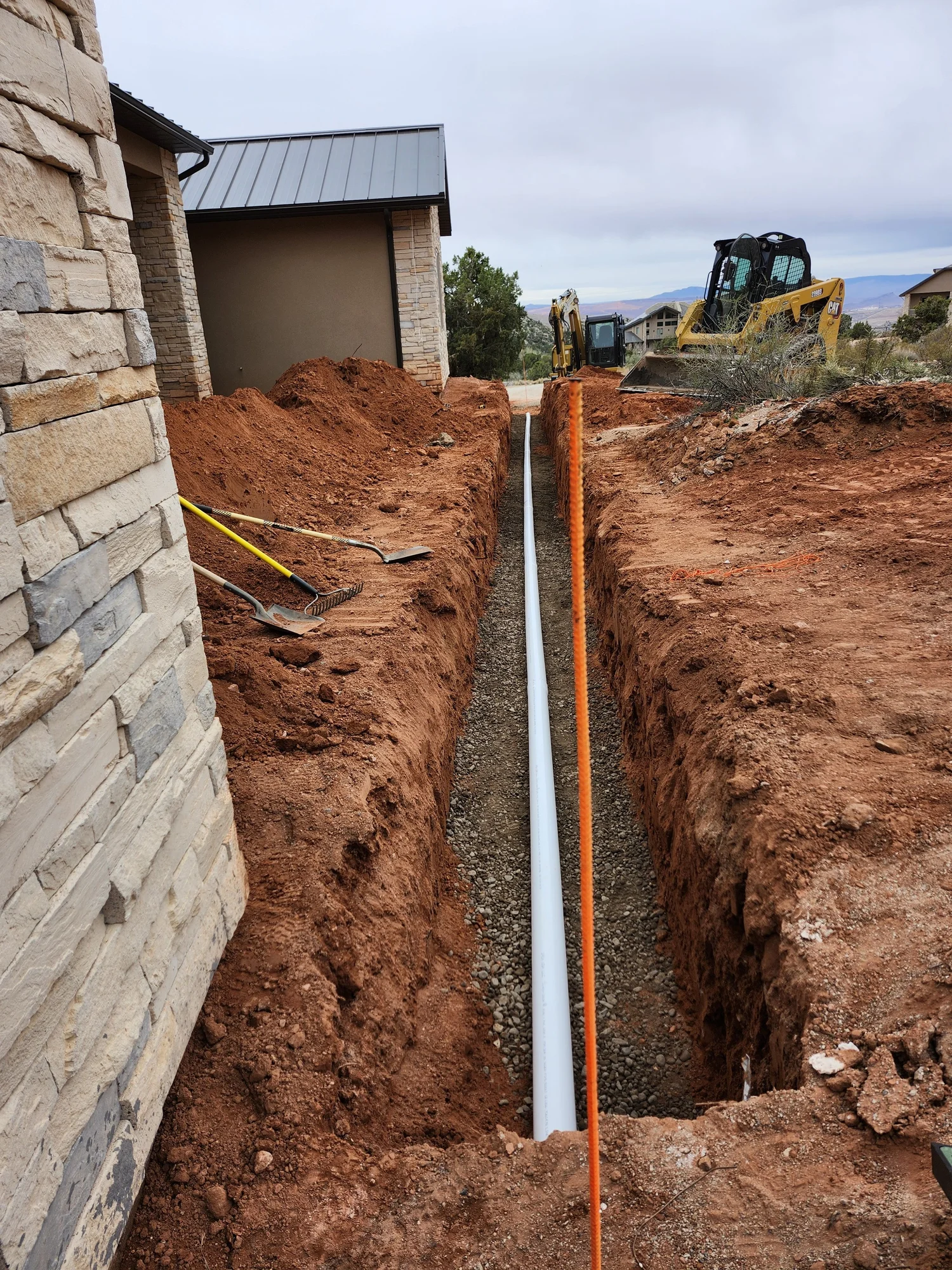 Underground white piping positioned in a prepared trench beside a stone-clad building, with Artisan Group excavation equipment visible