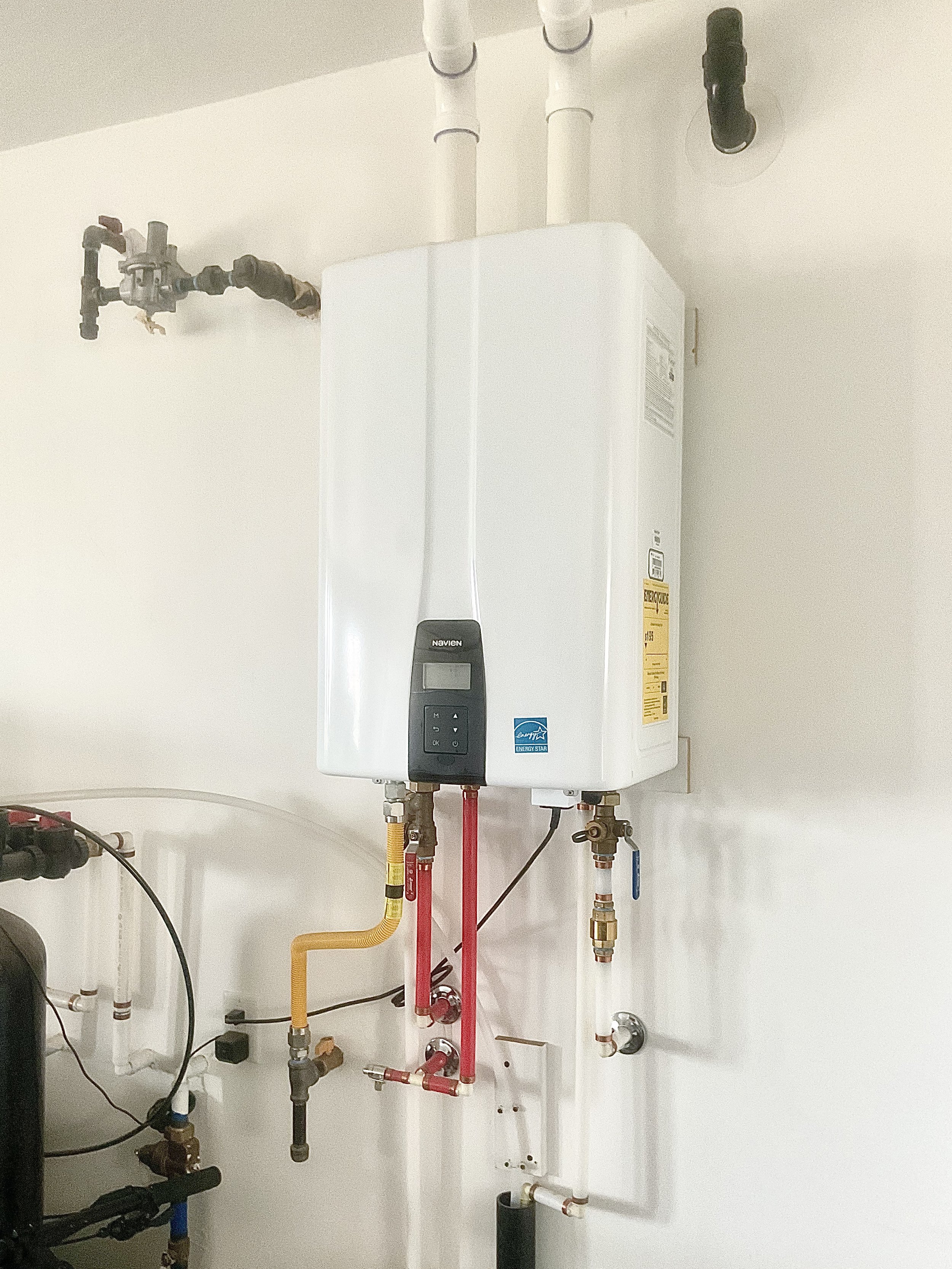 White tankless water heater with control panel, connected to various pipes and valves on a white wall.