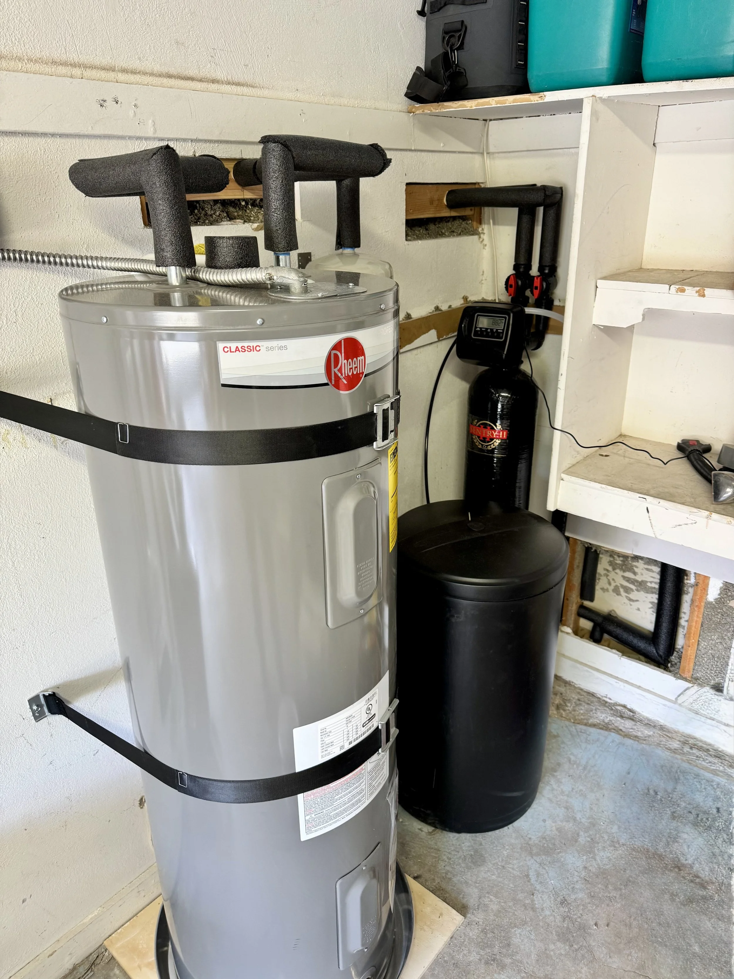 A water heater with black foam pipe insulation, a black water softener tank, and some black pipes on a wall in a laundry or utility room.