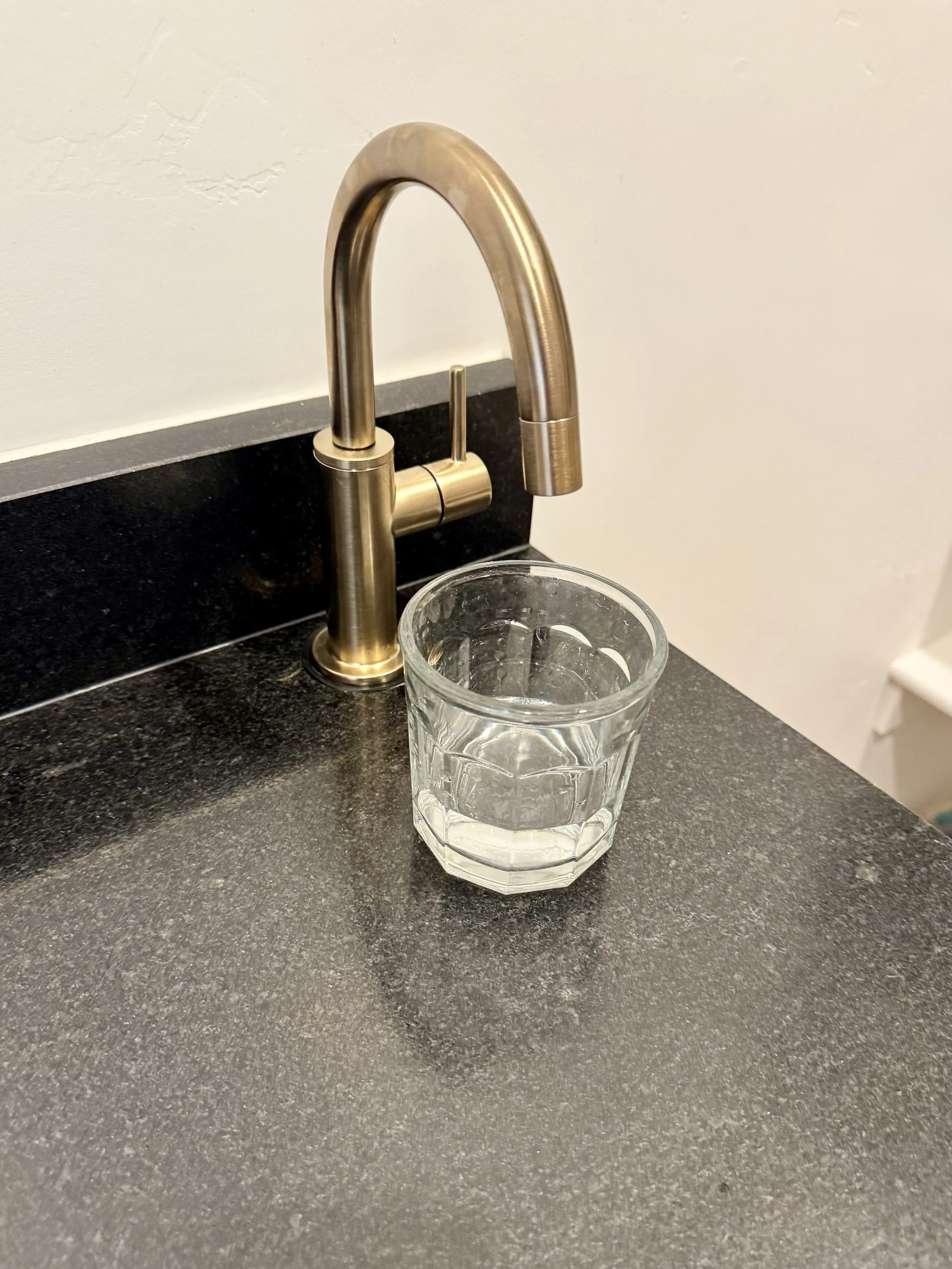 A kitchen sink with a modern stainless steel faucet and a clear glass of water on a black granite countertop.