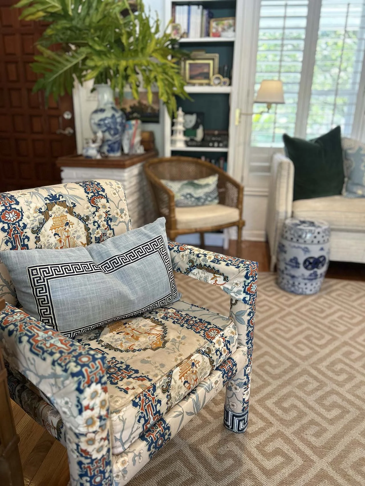 A living room with a patterned upholstered armchair with a rectangular pillow, a wicker chair with a pillow, and a white sofa with two pillows. There is a large blue and white vase, a bookshelf, and a window with blinds in the background.