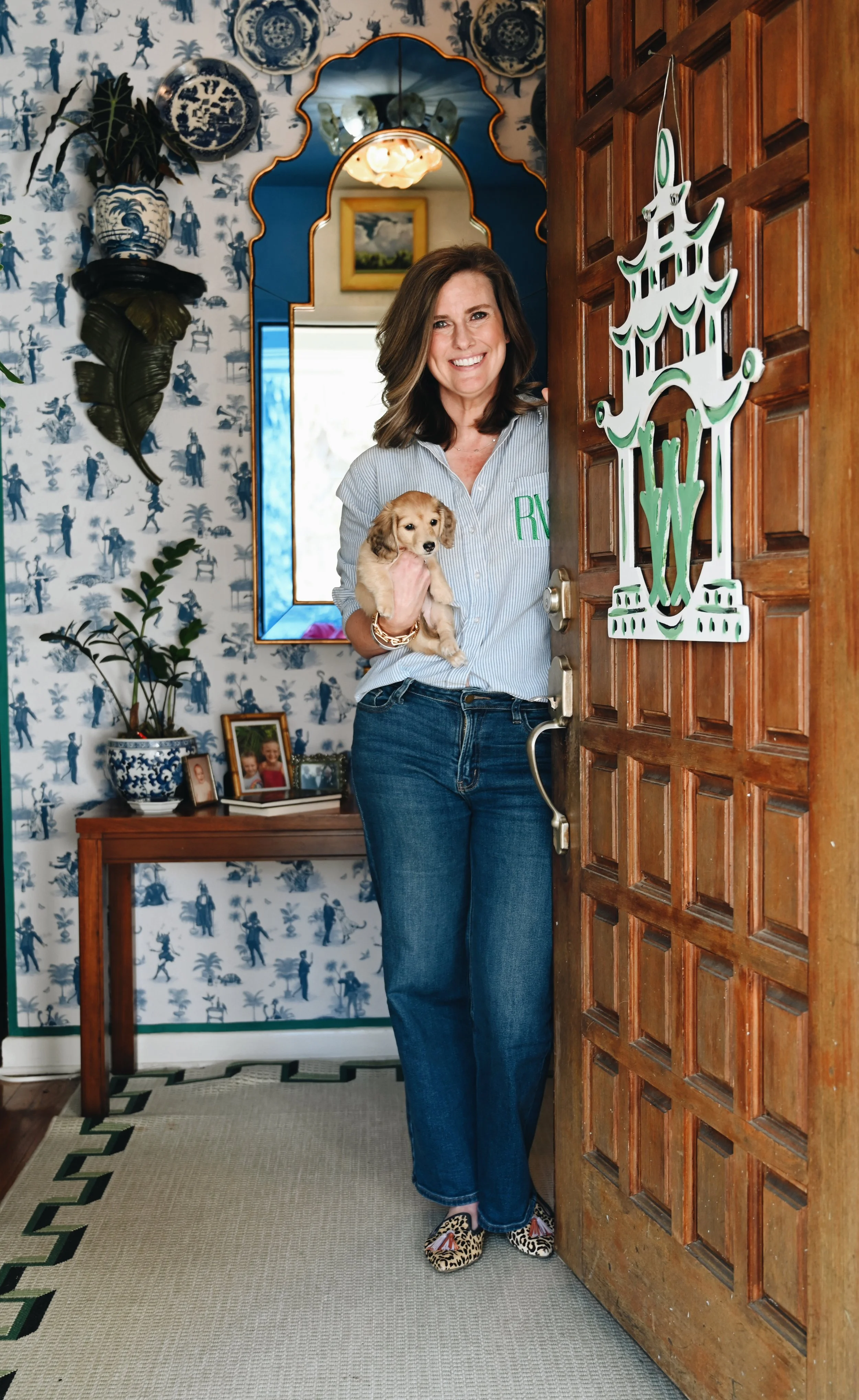 A woman smiling and holding a puppy, standing at the door of a home decorated with blue and white patterned wallpaper, framed photos on a console table, and a large mirror.