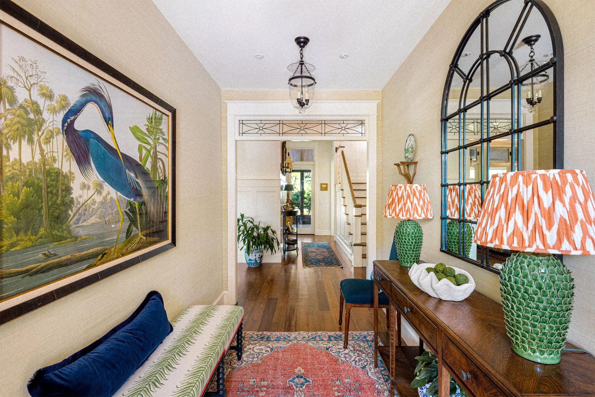 Entryway with hardwood floors, a gallery rug, and a wooden console table with green vases and orange-patterned lamps. A large framed artwork of a heron is on the left wall. There is a mirror above the console table and a staircase at the back.