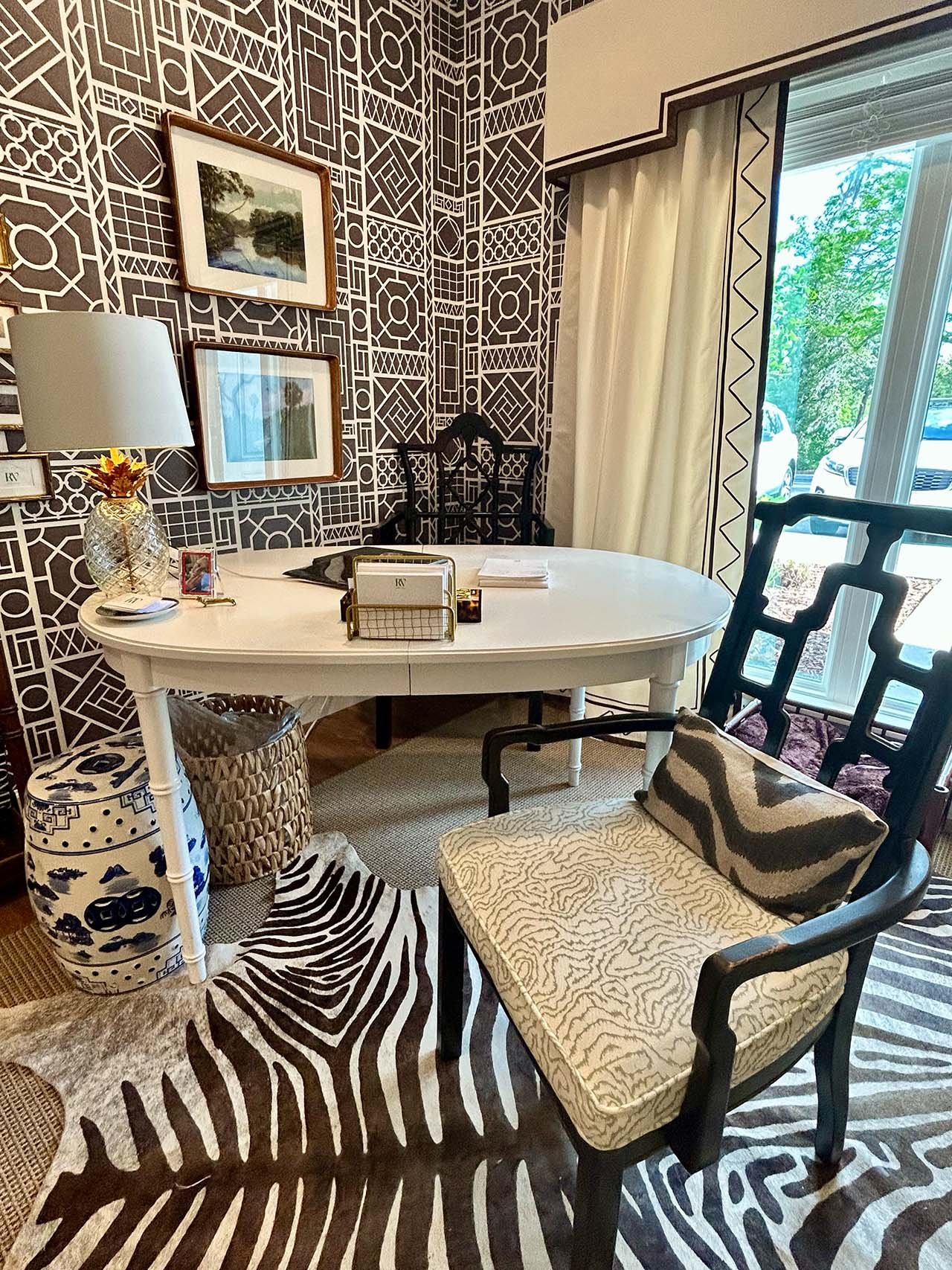 A stylish home office with a patterned black and white wallpaper, a white oval desk, chairs with animal print cushions, framed artwork on the wall, a table lamp, and a window with cream curtains showing parked cars outside.