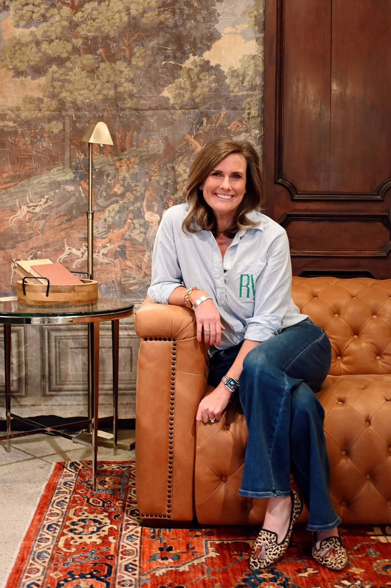 A woman with shoulder-length brown hair sitting on a tan leather tufted sofa, smiling, in a room with wood paneling and a large nature-themed mural in the background. She is wearing a striped button-up shirt with embroidered initials, jeans, and leopard-print high heels. There is a side table with a lamp and a wooden tray holding papers.