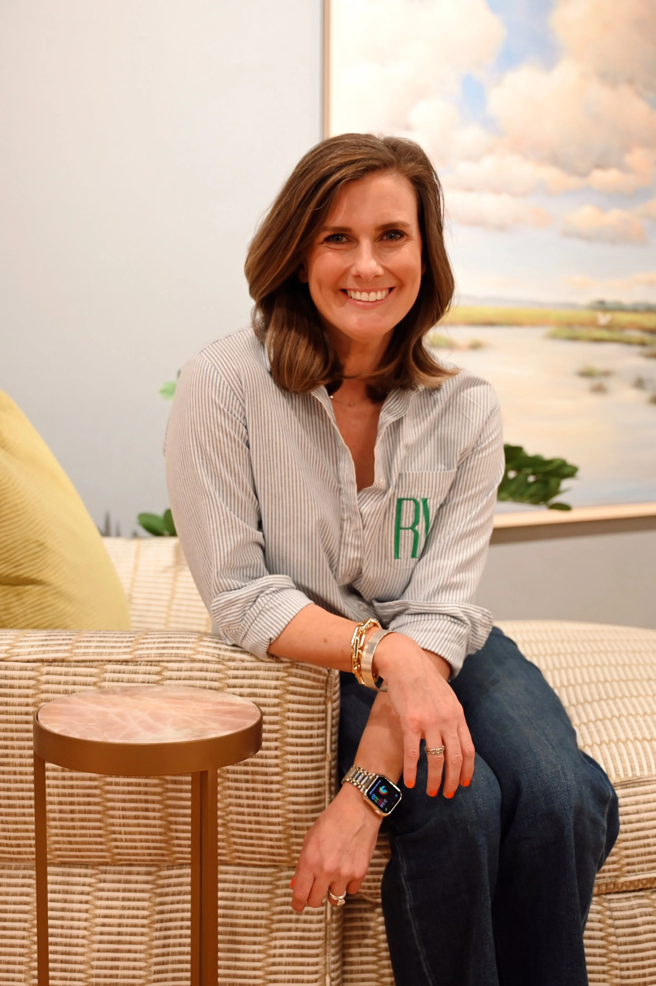 A woman with shoulder-length brown hair, smiling, wearing a striped button-up shirt with monogram initials, dark jeans, and a smartwatch, sitting on a woven beige sofa next to a small round side table in front of a landscape painting with clouds.