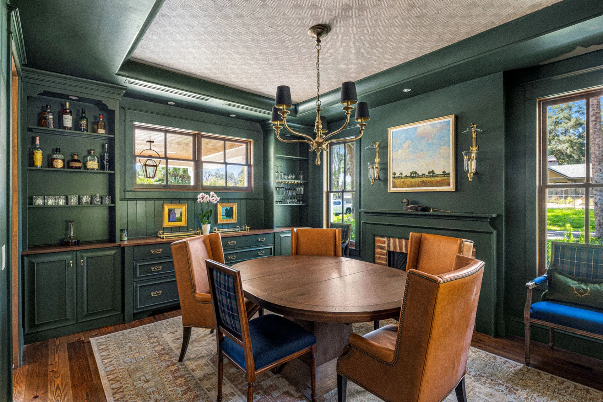 A dining room with dark green walls, a wooden table surrounded by six chairs with leather and plaid upholstery, a chandelier hanging above, a fireplace decorated with artwork and brass sconces, a built-in bar with shelves of liquor bottles and glasses, large windows showing trees and a lawn outside, and a patterned rug on wooden flooring.