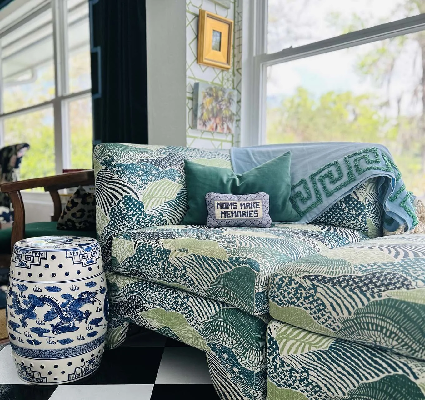 Living room with a patterned sofa and pillows, including one with the text 'MOMS MAKE MEMORIES.' A blue and white ceramic side table with dragon design, large windows with a view of trees outside, and artwork on the wall.