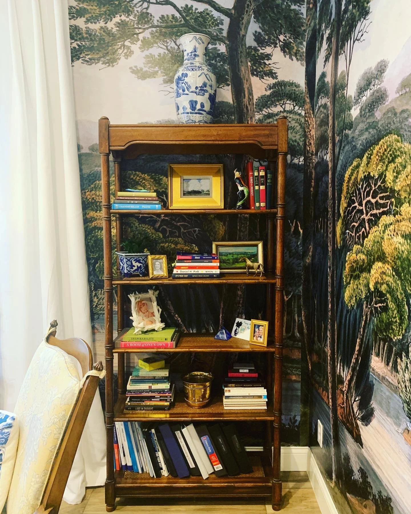 A wooden bookshelf filled with books, framed photos, and decorative items, standing against a mural of trees and nature on the wall.