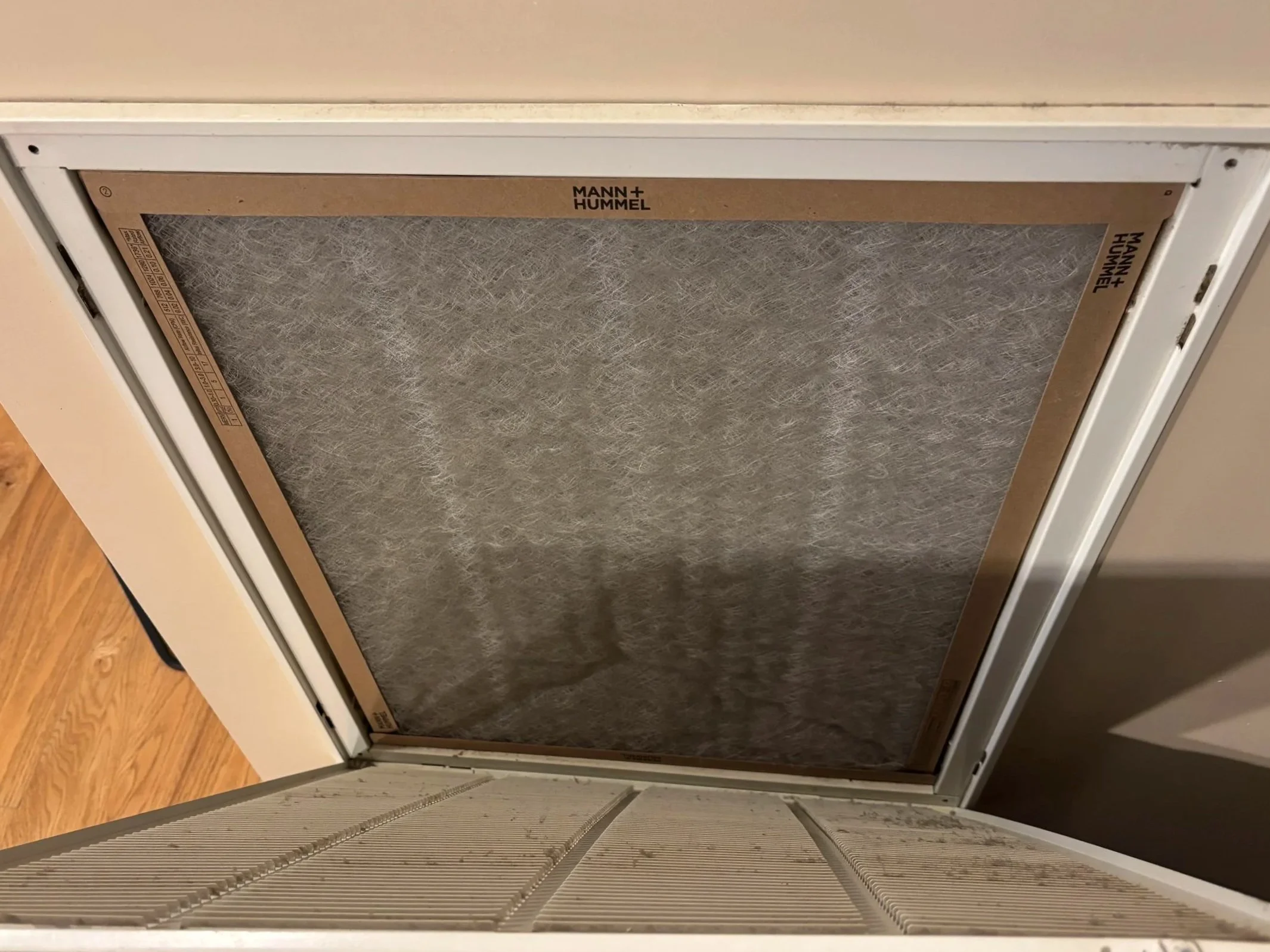 An HVAC return-air grille opened to reveal a residential air filter during a routine inspection. The filter shows normal dust accumulation and is installed inside a ceiling or wall-mounted return vent.