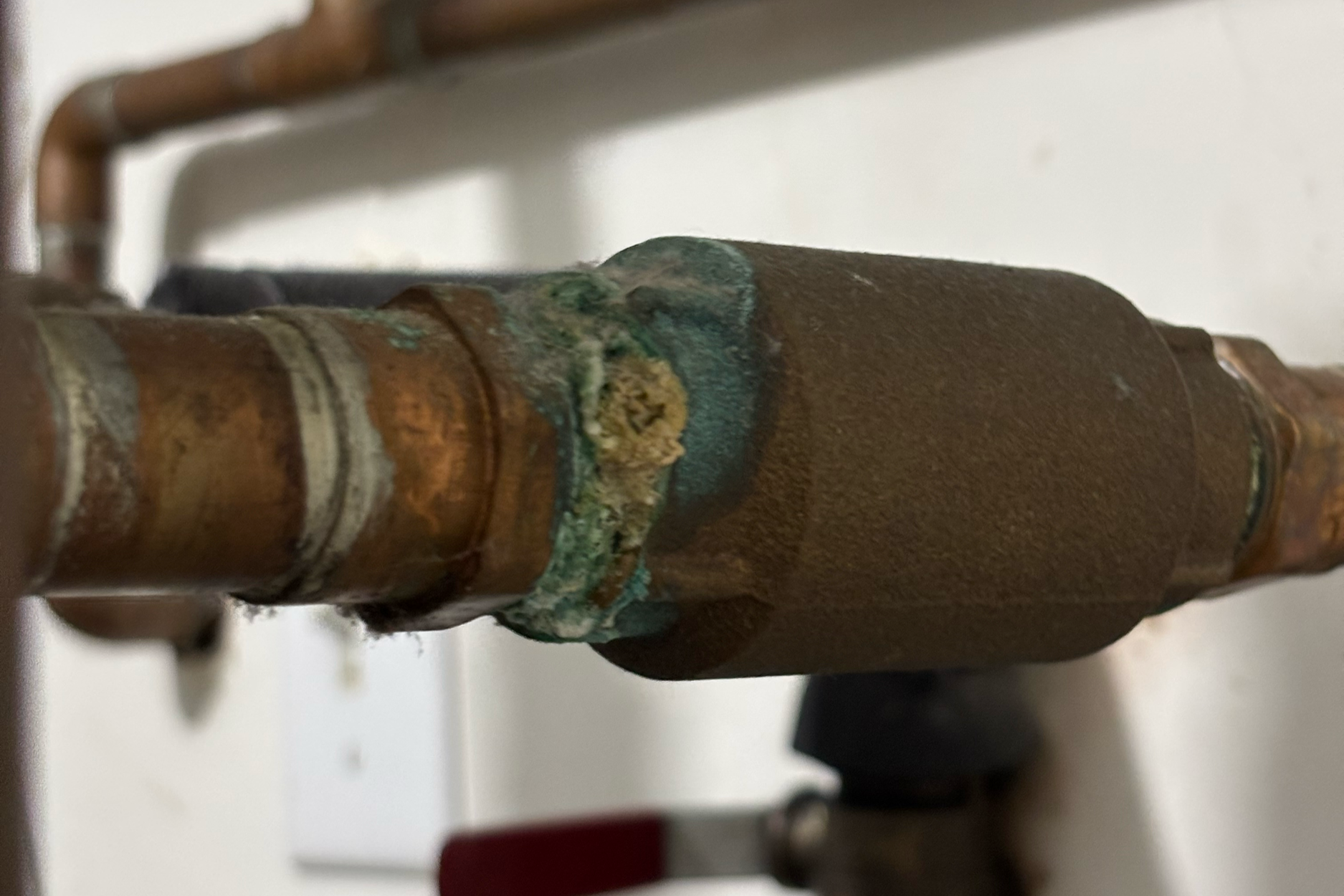 A close-up photograph of an inline check valve installed on a copper water supply line showing visible signs of deterioration and stress.
