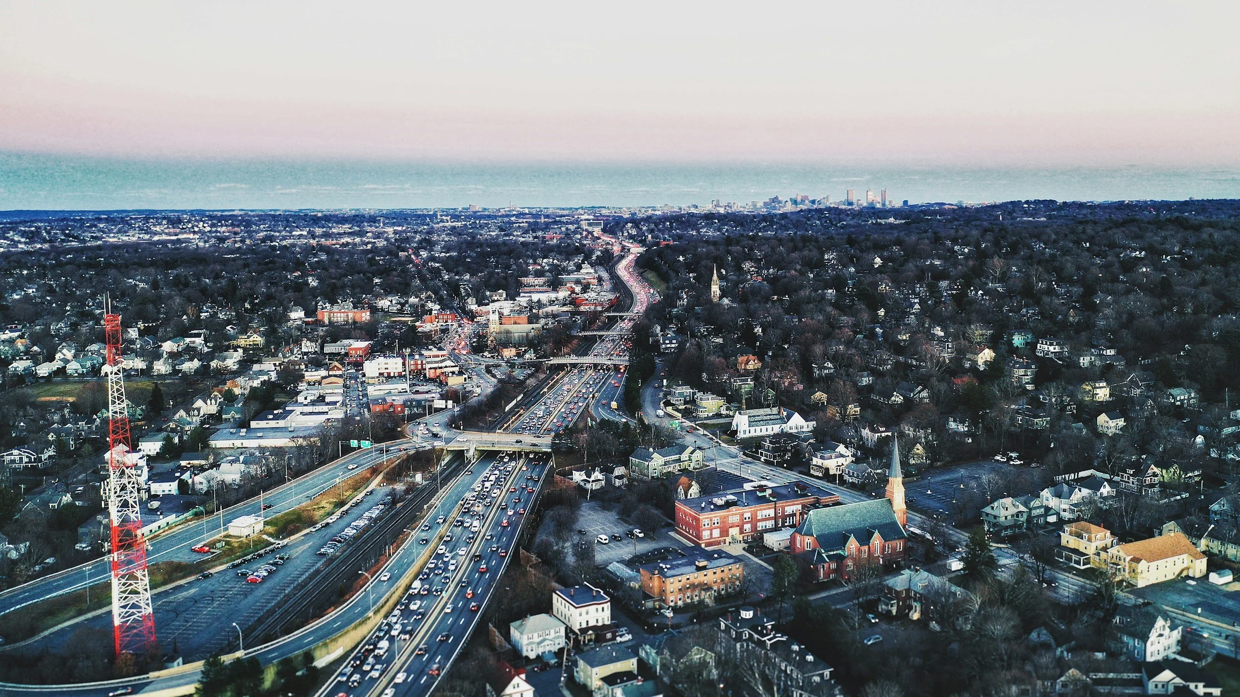 An aerial view of a cityscape with a busy highway filled with cars, residential houses, and a church, leading towards a distant downtown skyline with high-rise buildings under a clear sky.