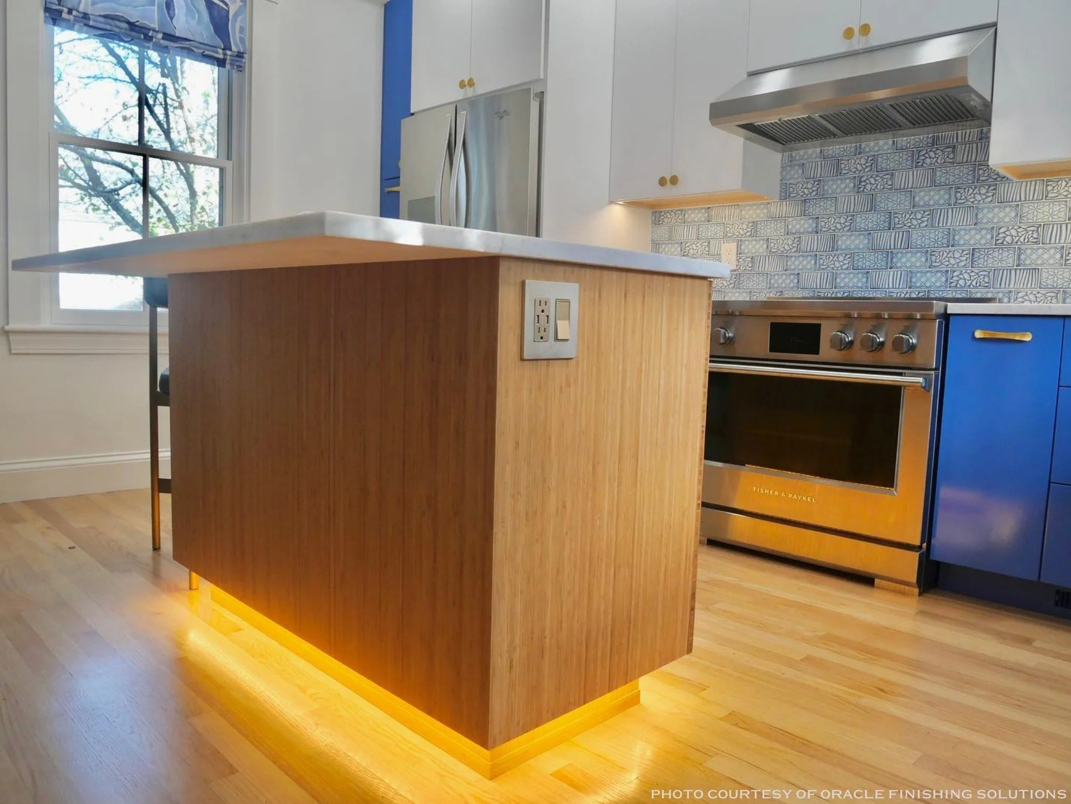 A modern kitchen with a wooden island with warm under-lighting, a window showing trees outside, white upper cabinets, a patterned tile backsplash, and stainless steel appliances including a refrigerator and oven.