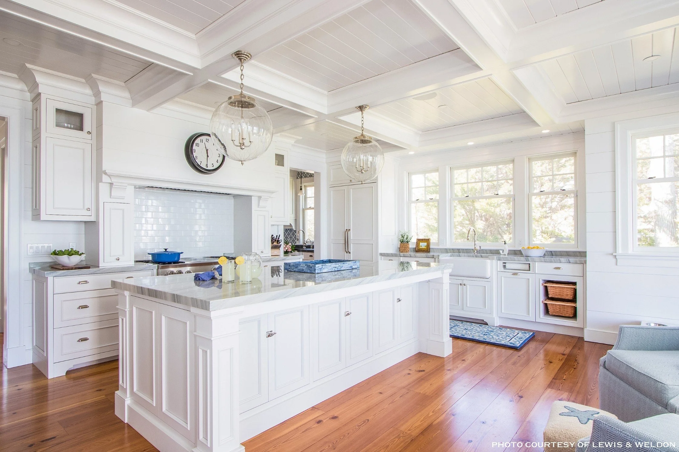 Bright white kitchen with a large island, white cabinetry, and decorative crown molding. Hanging glass pendant lights, a wall clock, and large windows letting in natural light.