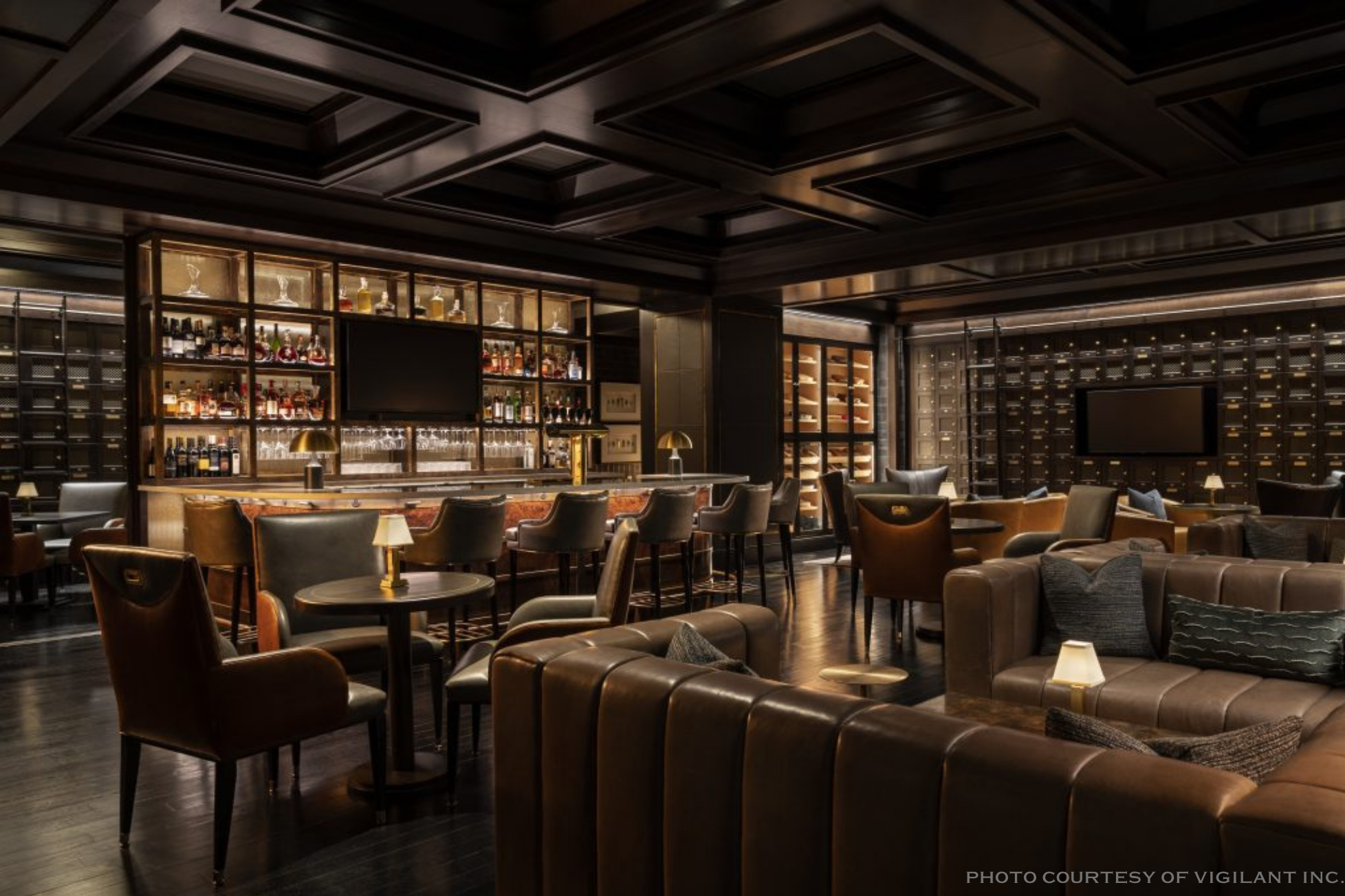 Luxurious bar and lounge with dark wood ceiling, shelves of alcohol bottles, large flat-screen TV, and plush leather seating with small table lamps.
