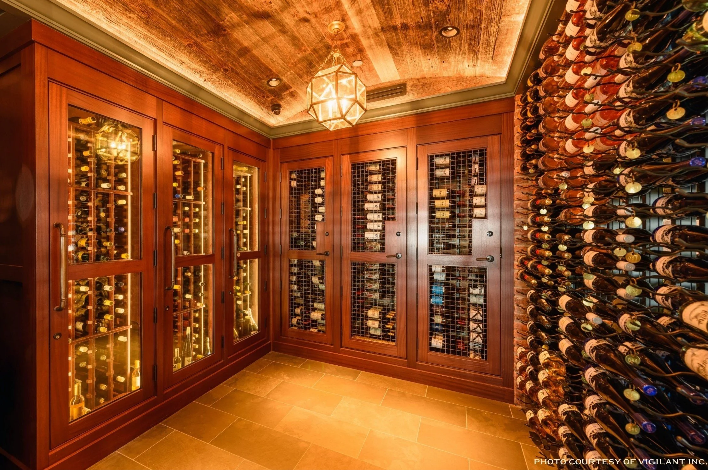A wine cellar with wooden cabinets and glass doors, shelves filled with wine bottles, a brick wall, and a wooden ceiling with a geometric chandelier.