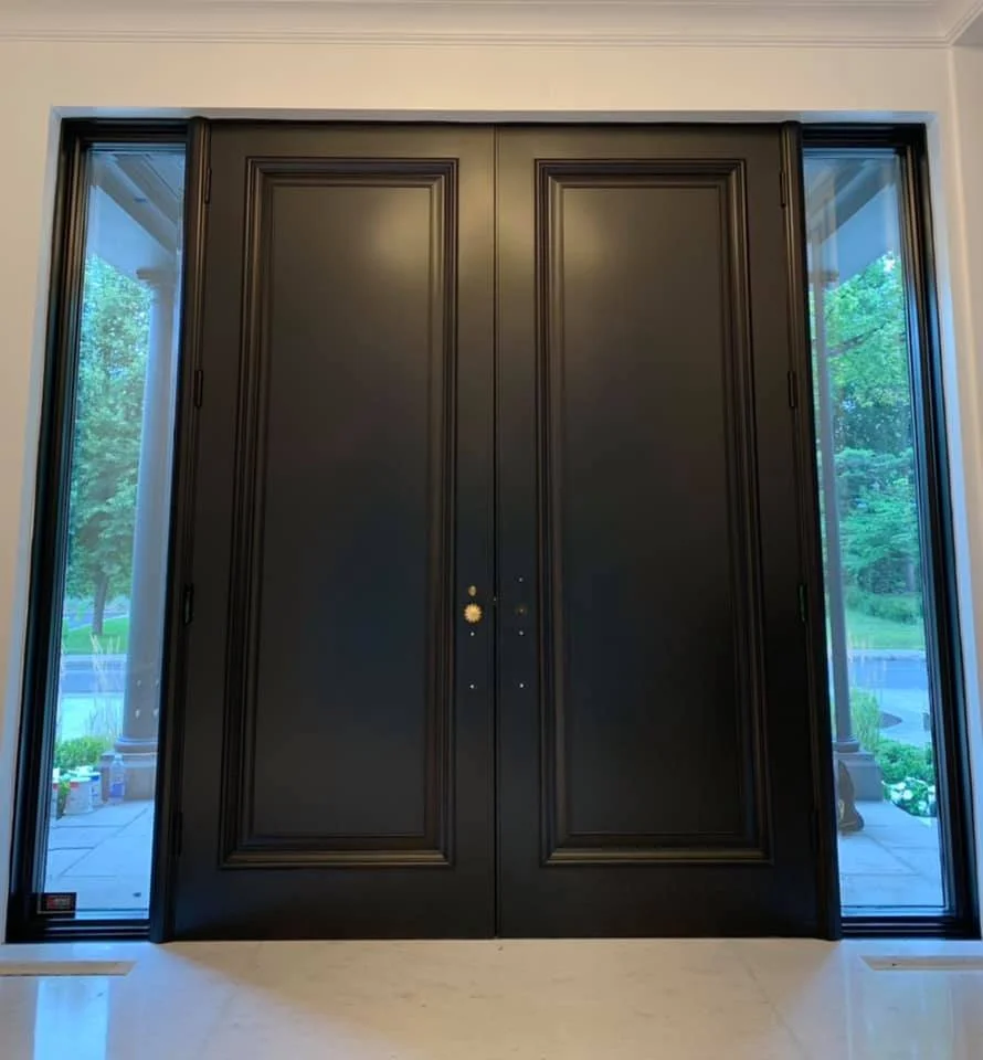 Black double doors with gold handles, flanked by glass panels showing an outdoor garden and patio area.