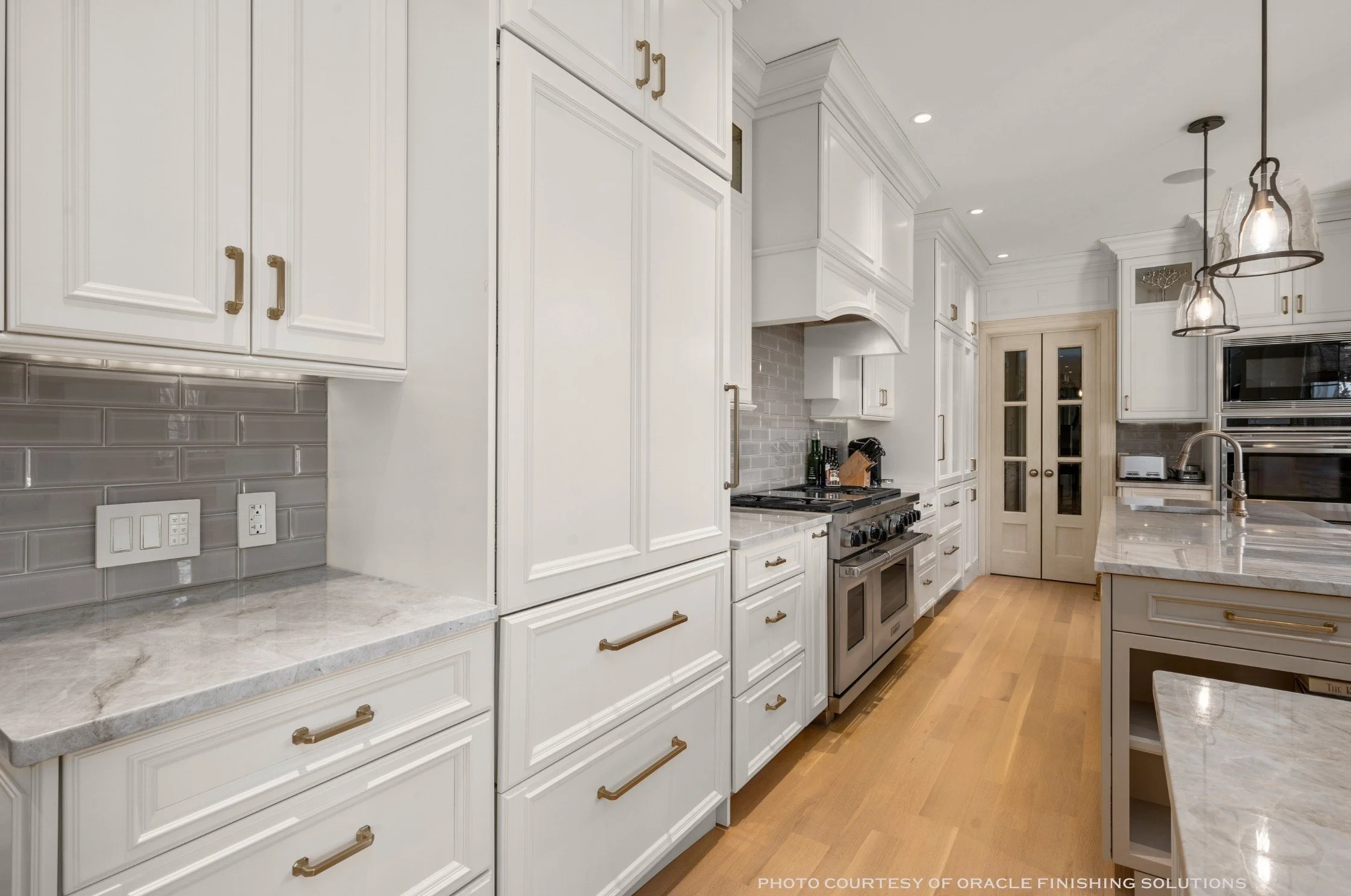 A modern kitchen with white cabinetry, a marble countertop, gray subway tile backsplash, and stainless steel appliances. There are pendant lights hanging above the kitchen island.